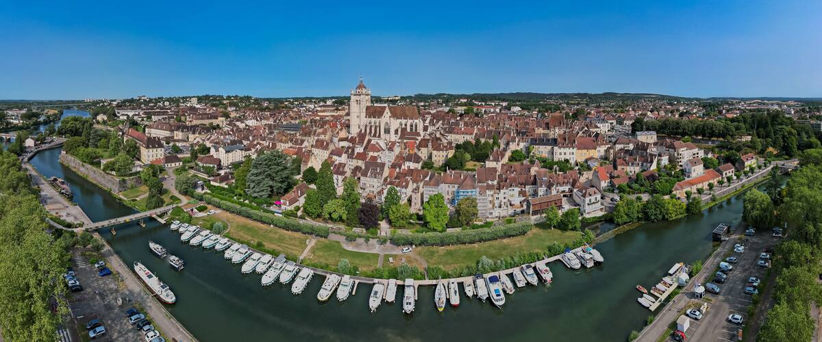 A river with boats docked along the banks, a historic town with a prominent church, and a bridge connecting two sides of the town.