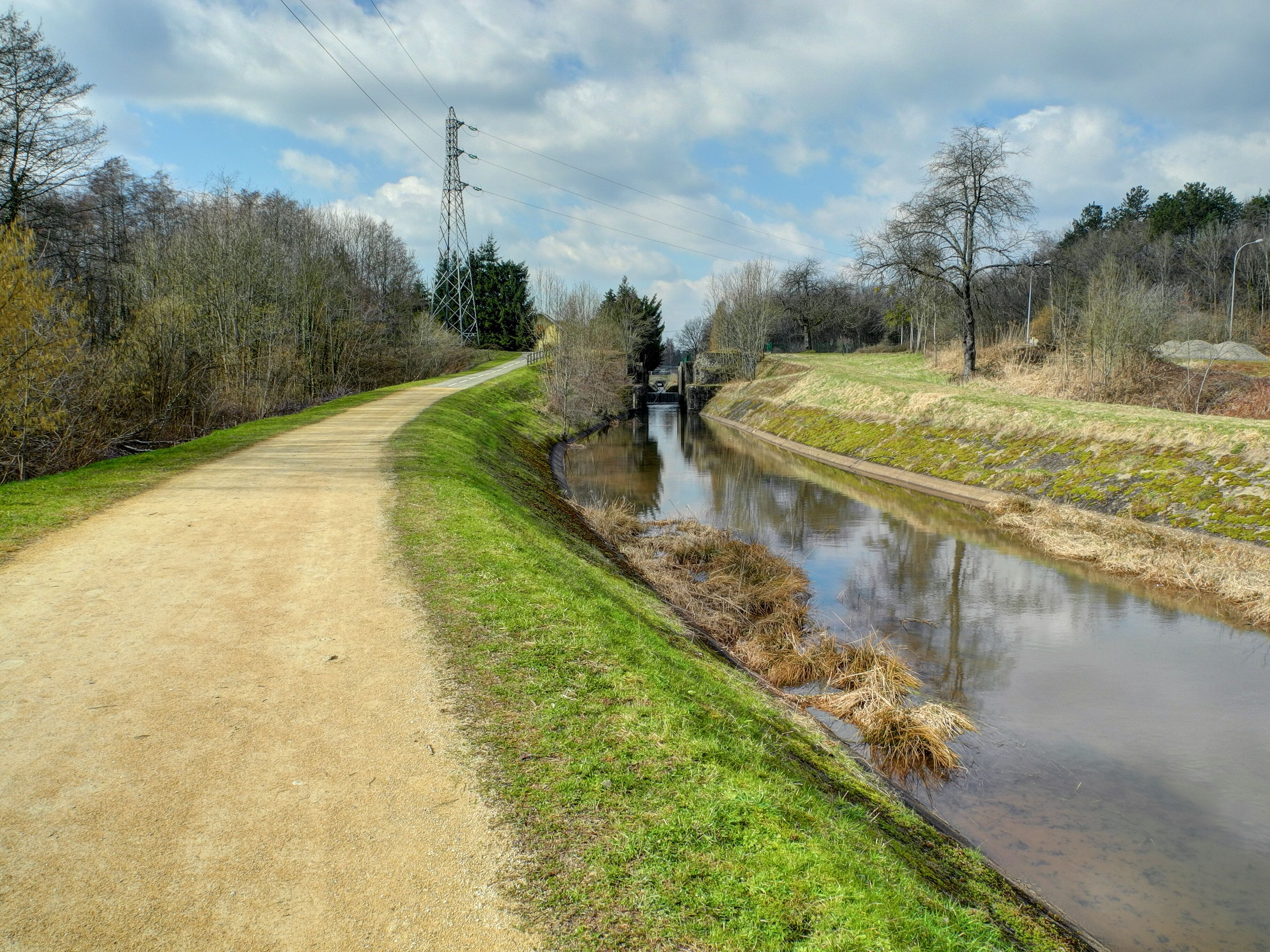 A dirt path beside a calm canal with trees and a power line tower in the background.