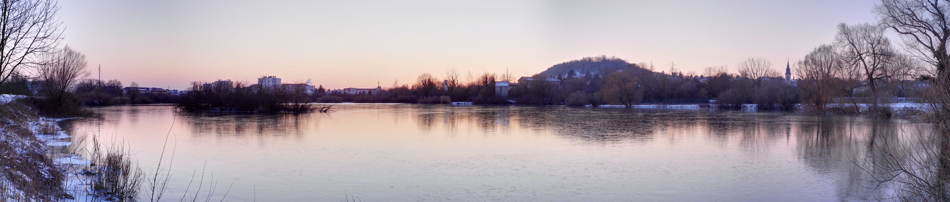 A calm river with reflections of trees and buildings, surrounded by snow-covered banks and a hill with a church spire in the background.