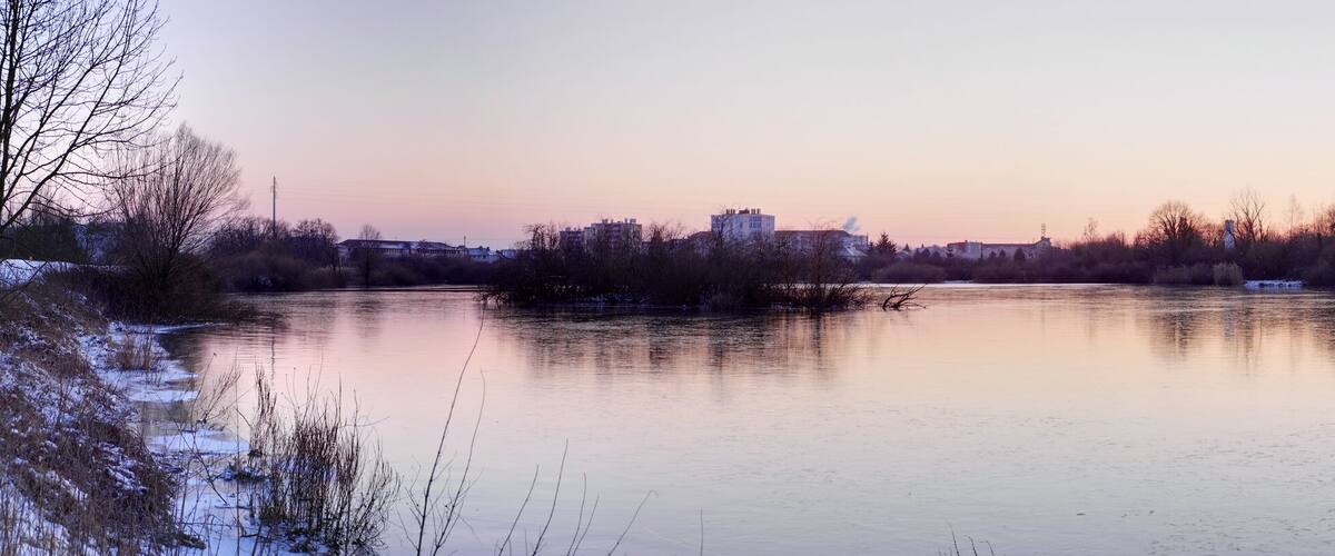 A calm river with reflections of trees and buildings, surrounded by snow-covered banks and a hill with a church spire in the background.
