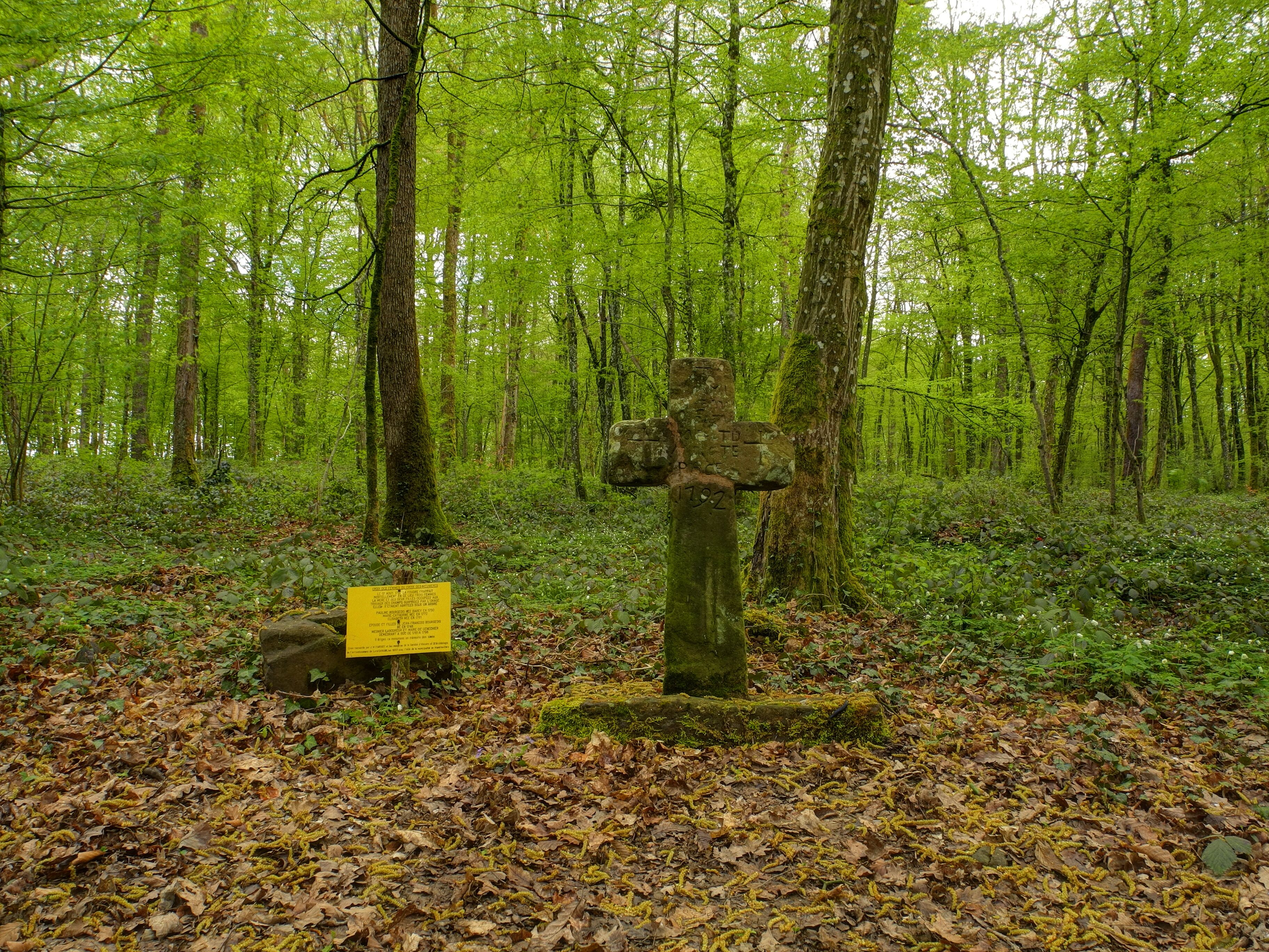 A stone cross in a forest with a yellow sign nearby.