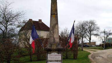 A war memorial with two French flags, surrounded by a circular stone pavement, with a house and trees in the background.