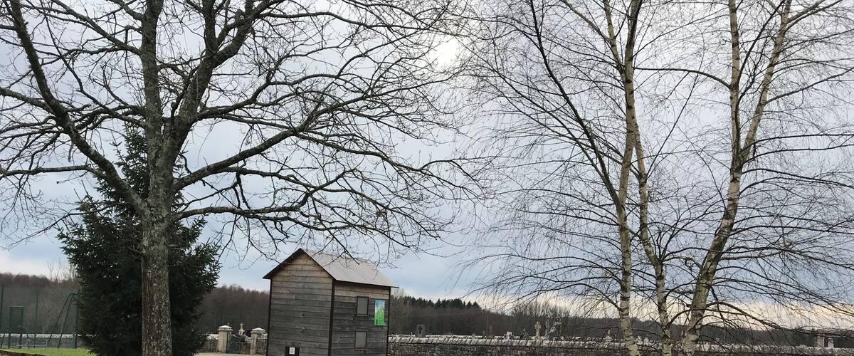 A small wooden structure in a grassy field with leafless trees and a stone wall in the background.