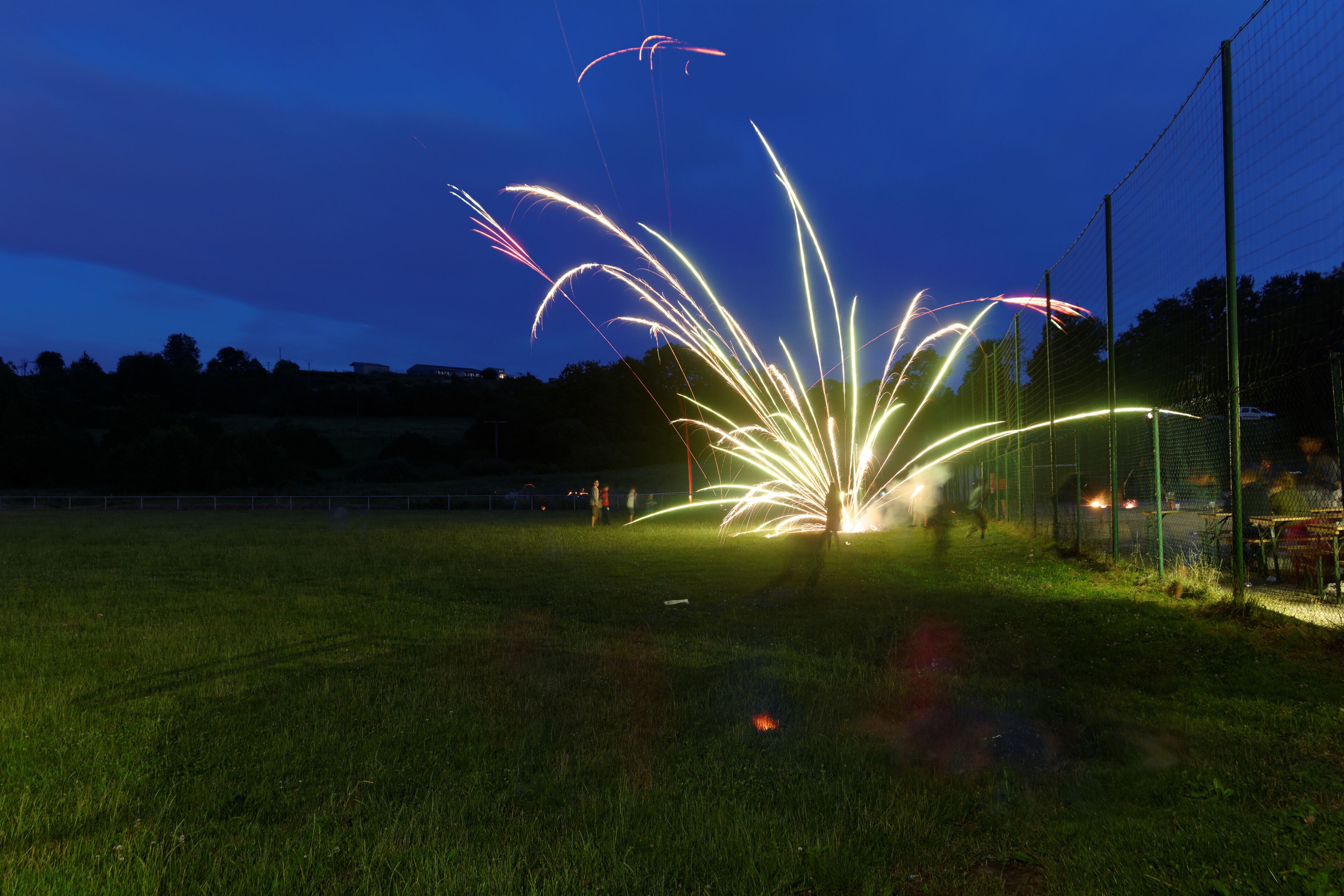 A nighttime scene with fireworks in a grassy field, a fence, and trees in the background.