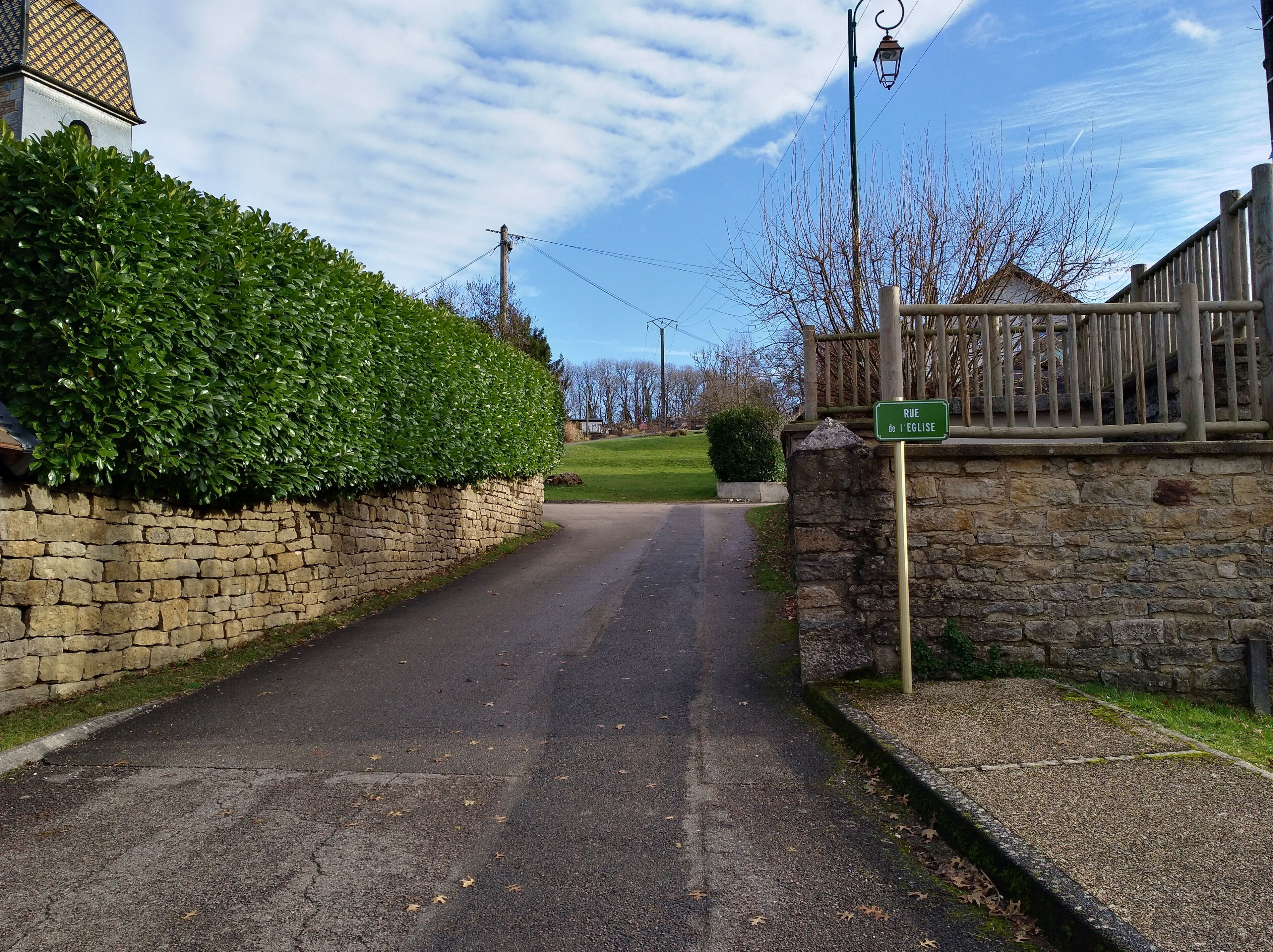 A rural road with a stone wall and a green hedge on one side, and a wooden fence on the other.