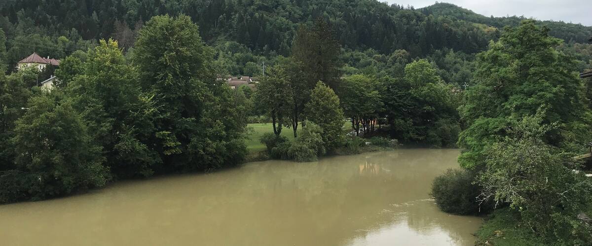 A river flowing through a valley with dense greenery and houses in the background.