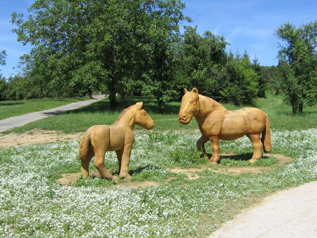 Two wooden horse statues in a grassy field with trees and a dirt path in the background.