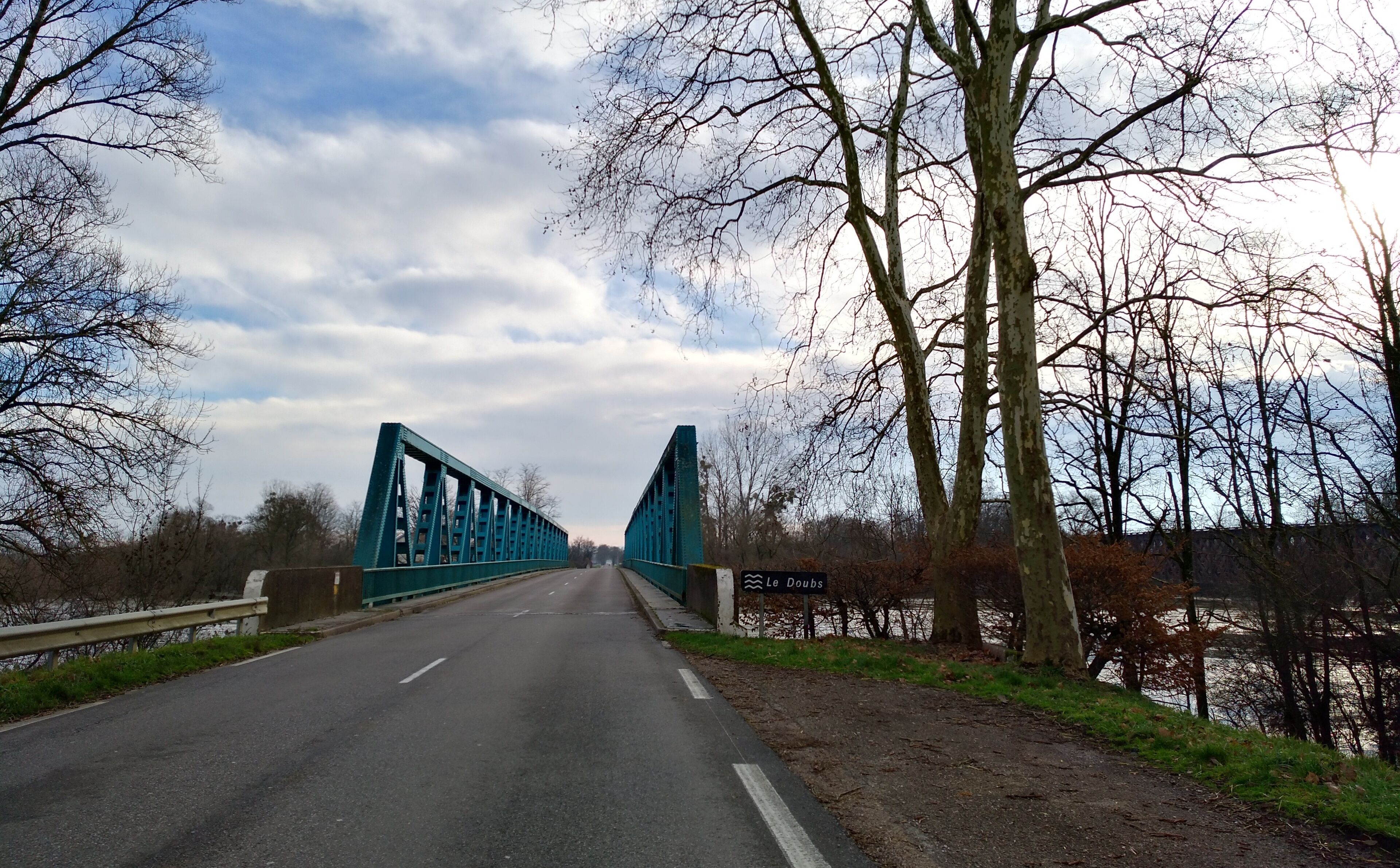A blue steel bridge over a road with a river on the right.