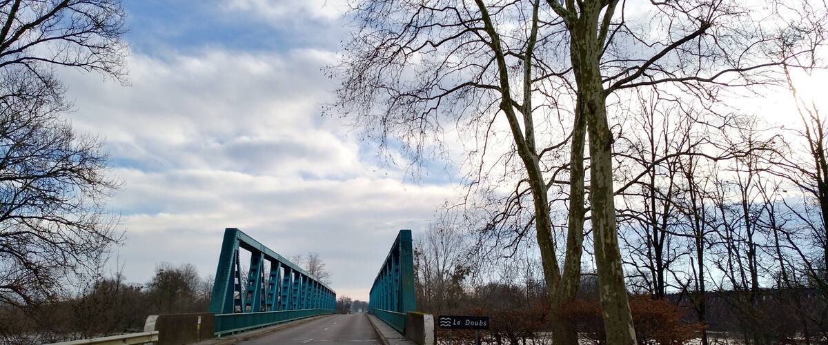 A blue steel bridge over a road with a river on the right.