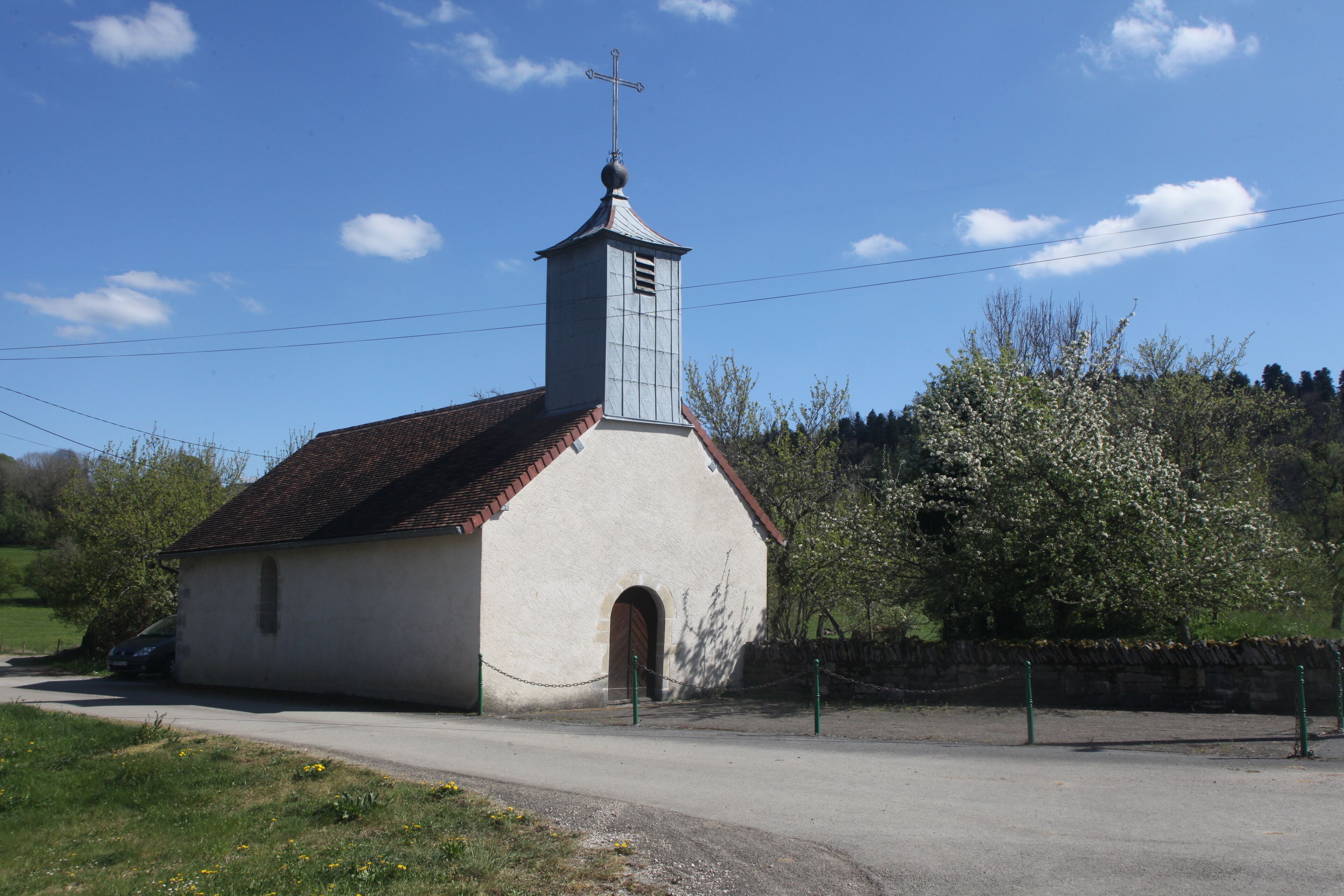 A small church with a bell tower and a cross on top, surrounded by trees and a stone wall.