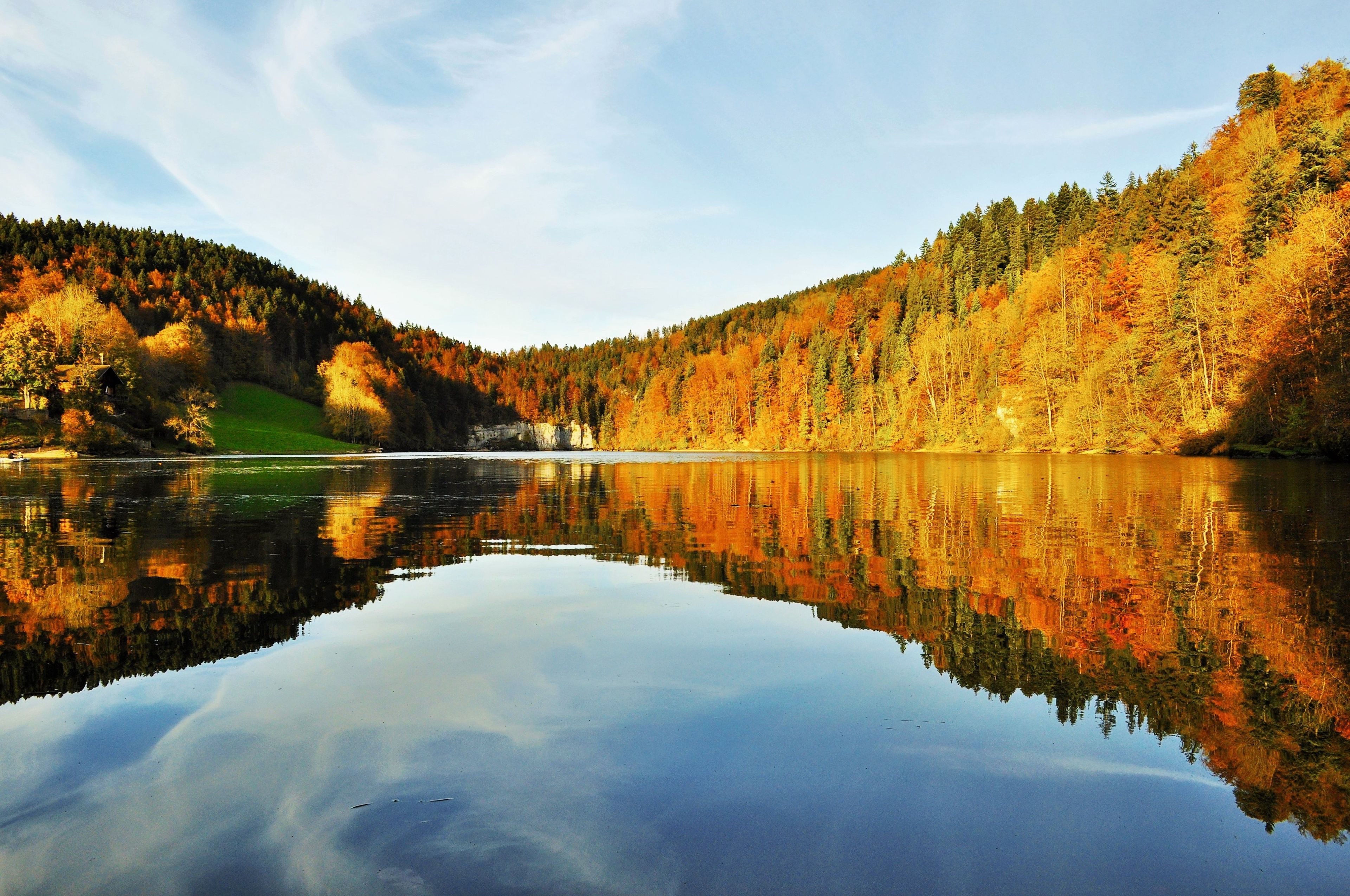 A calm lake reflecting autumn trees and a small cabin on a hillside.