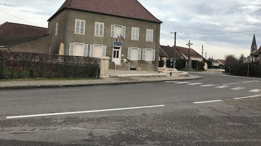 A two-story building with a red roof and white shutters, a sign reading "MAIRIE," and a church with a steeple in the background.