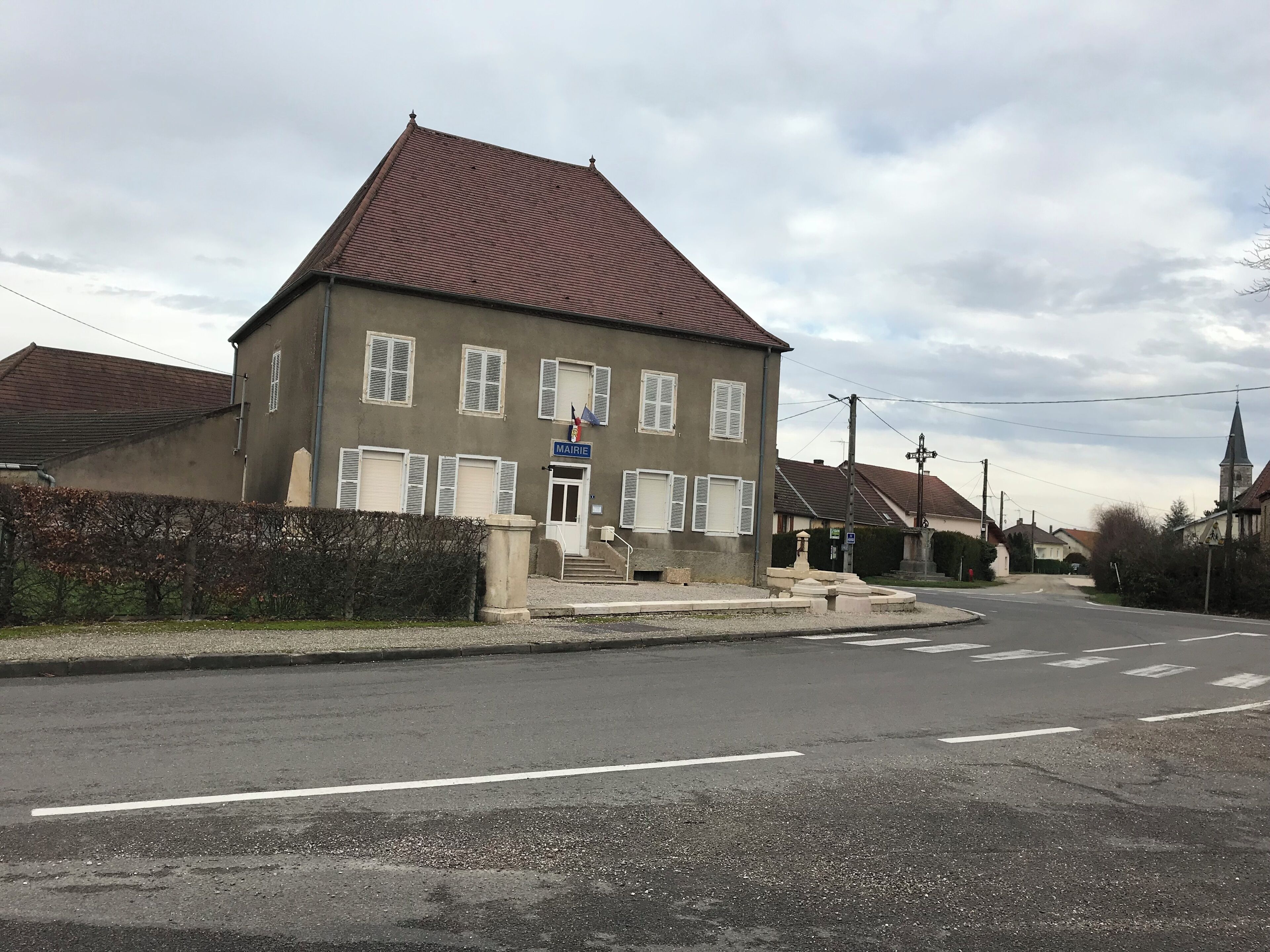 A two-story building with a red roof and white shutters, a sign reading "MAIRIE," and a church with a steeple in the background.