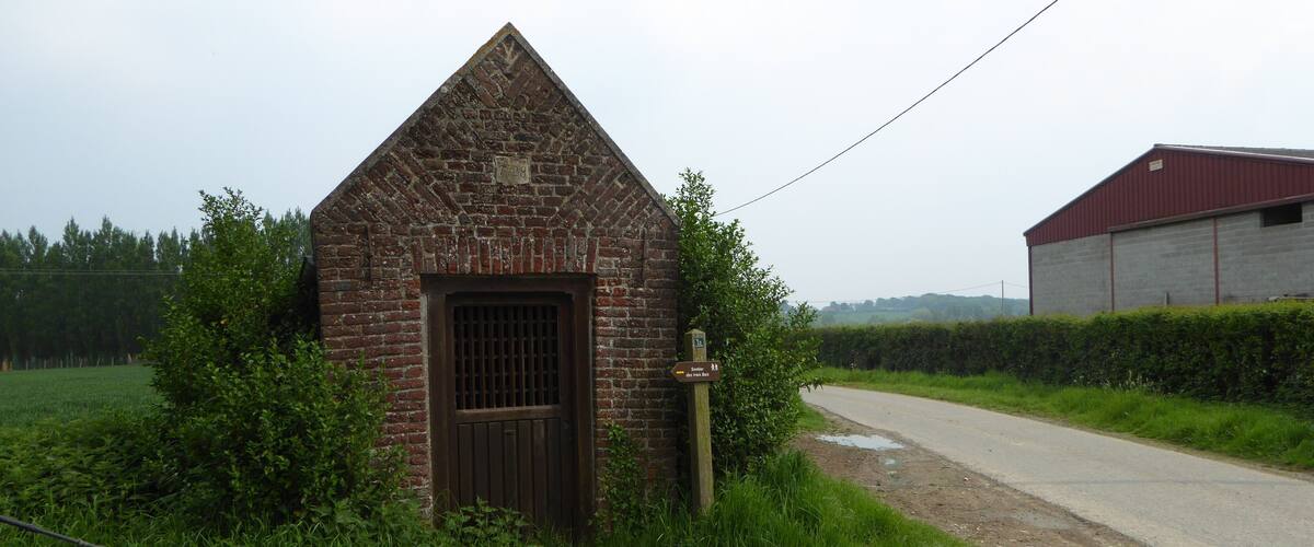 A small brick building with a pointed roof and a metal door, surrounded by lush greenery and a rural road.