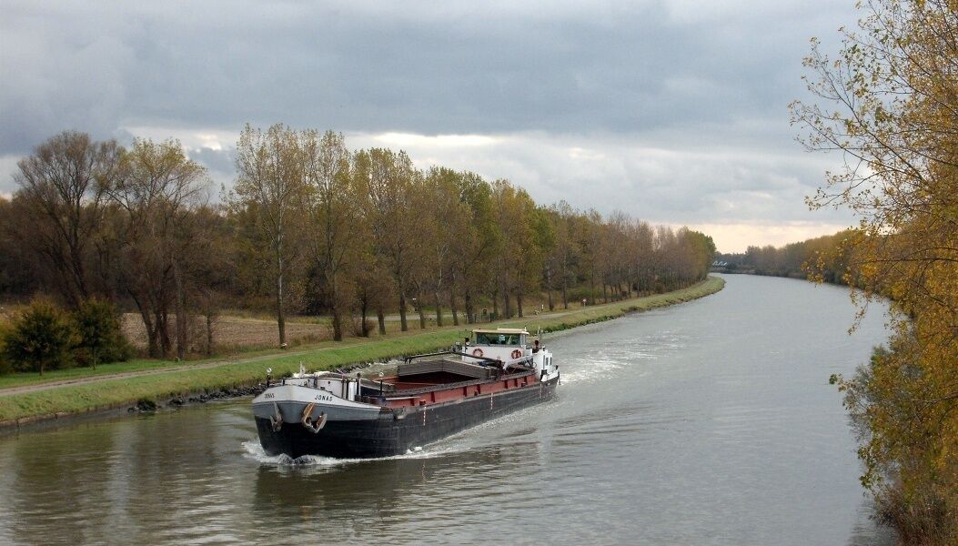 A river with a barge moving along it, surrounded by trees and a cloudy sky.