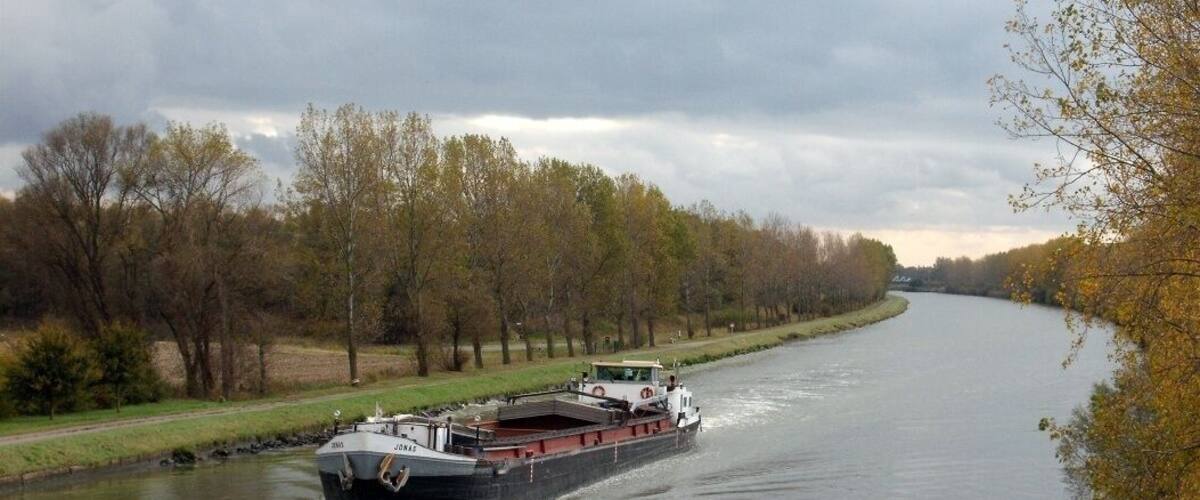 A river with a barge moving along it, surrounded by trees and a cloudy sky.