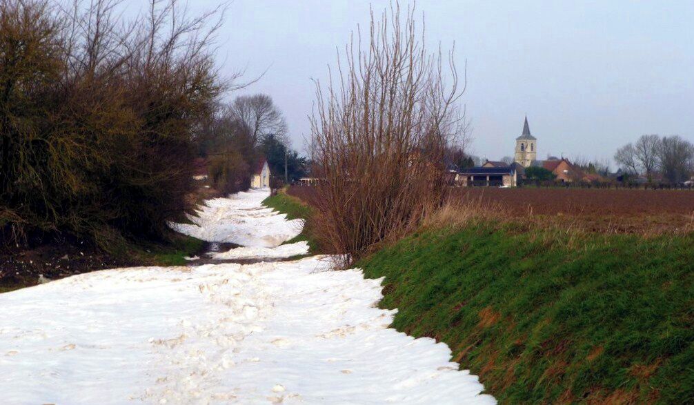 A snow-covered path leading to a church with a tower, surrounded by bare trees and fields.