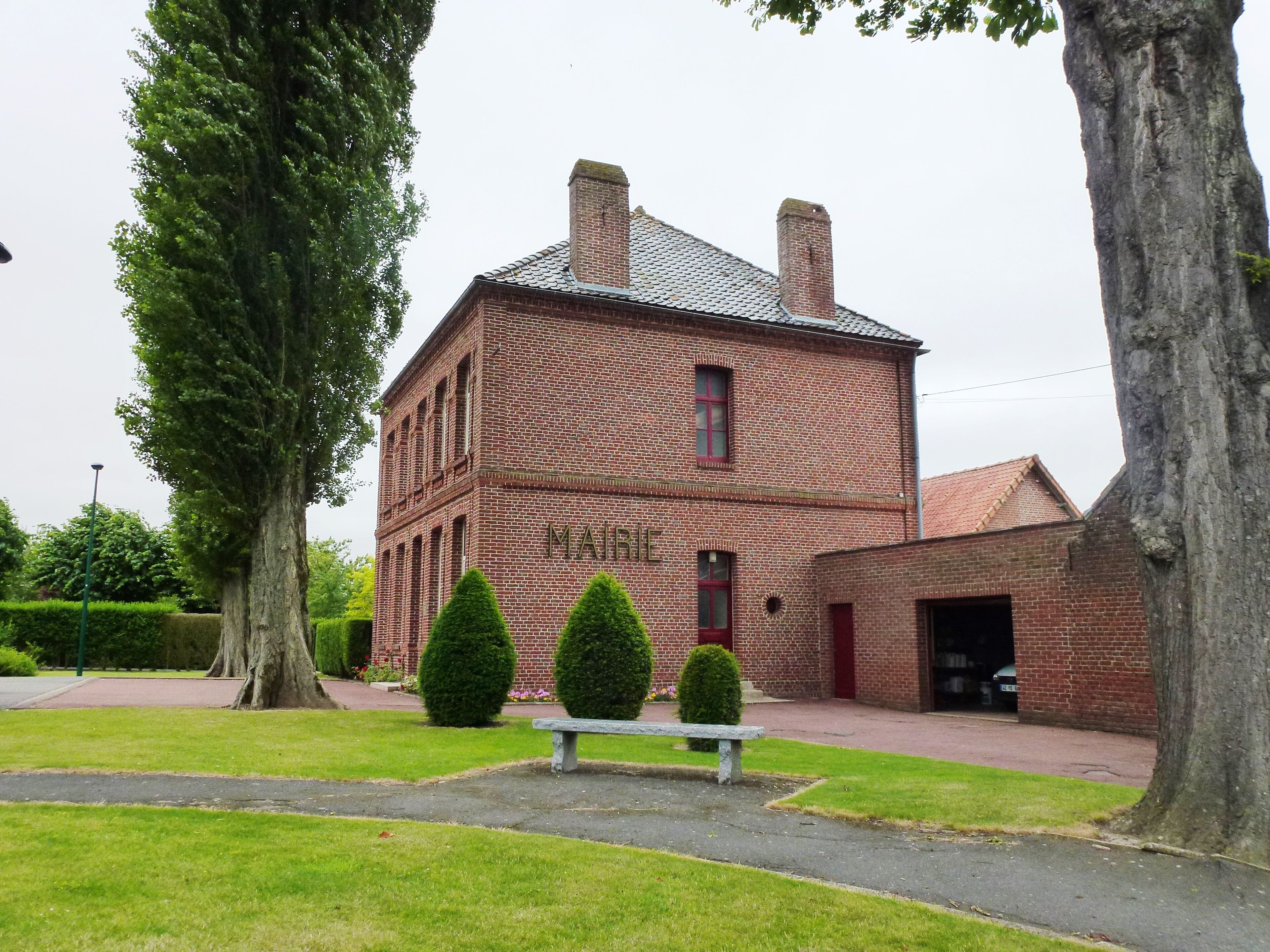 A brick building labeled "MAIRIE" with a red roof, surrounded by green grass, trees, and a bench.