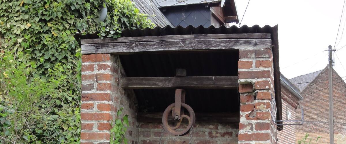 A brick structure with a metal ring pulley, surrounded by greenery and a brick building in the background.