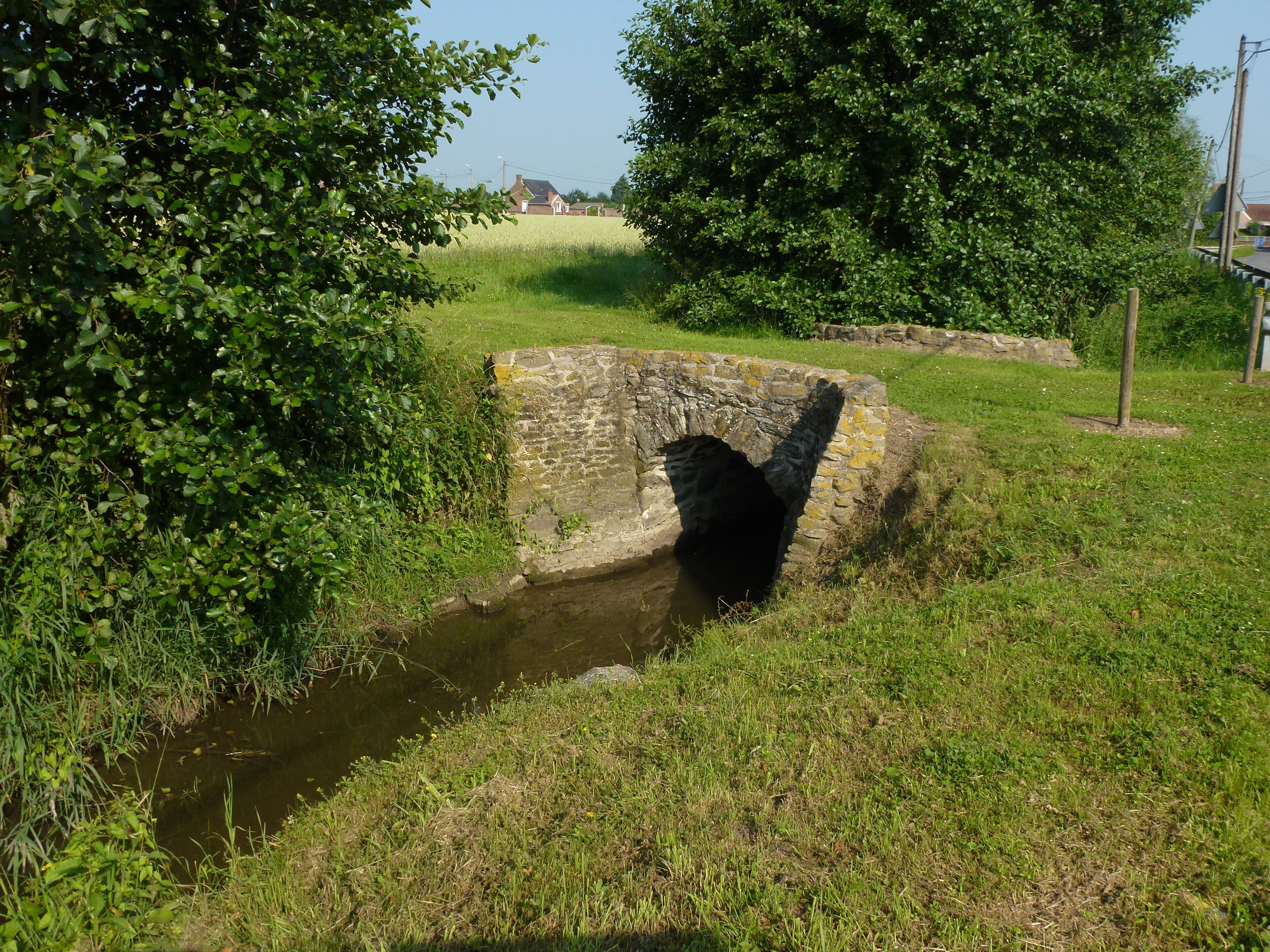 A stone bridge over a narrow water channel, surrounded by lush greenery and trees.