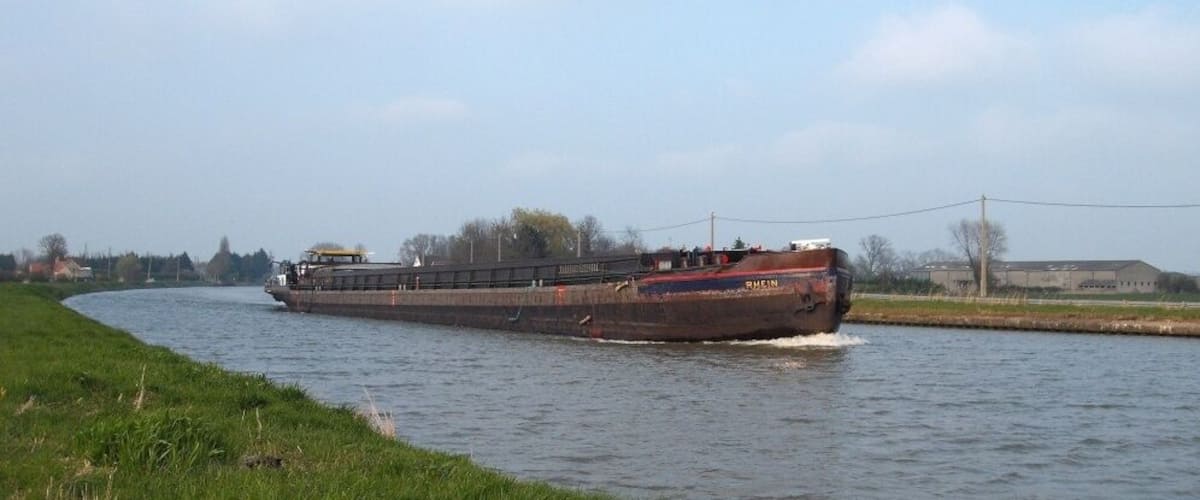 A large cargo ship moving through a narrow waterway with grassy banks and industrial buildings in the background.