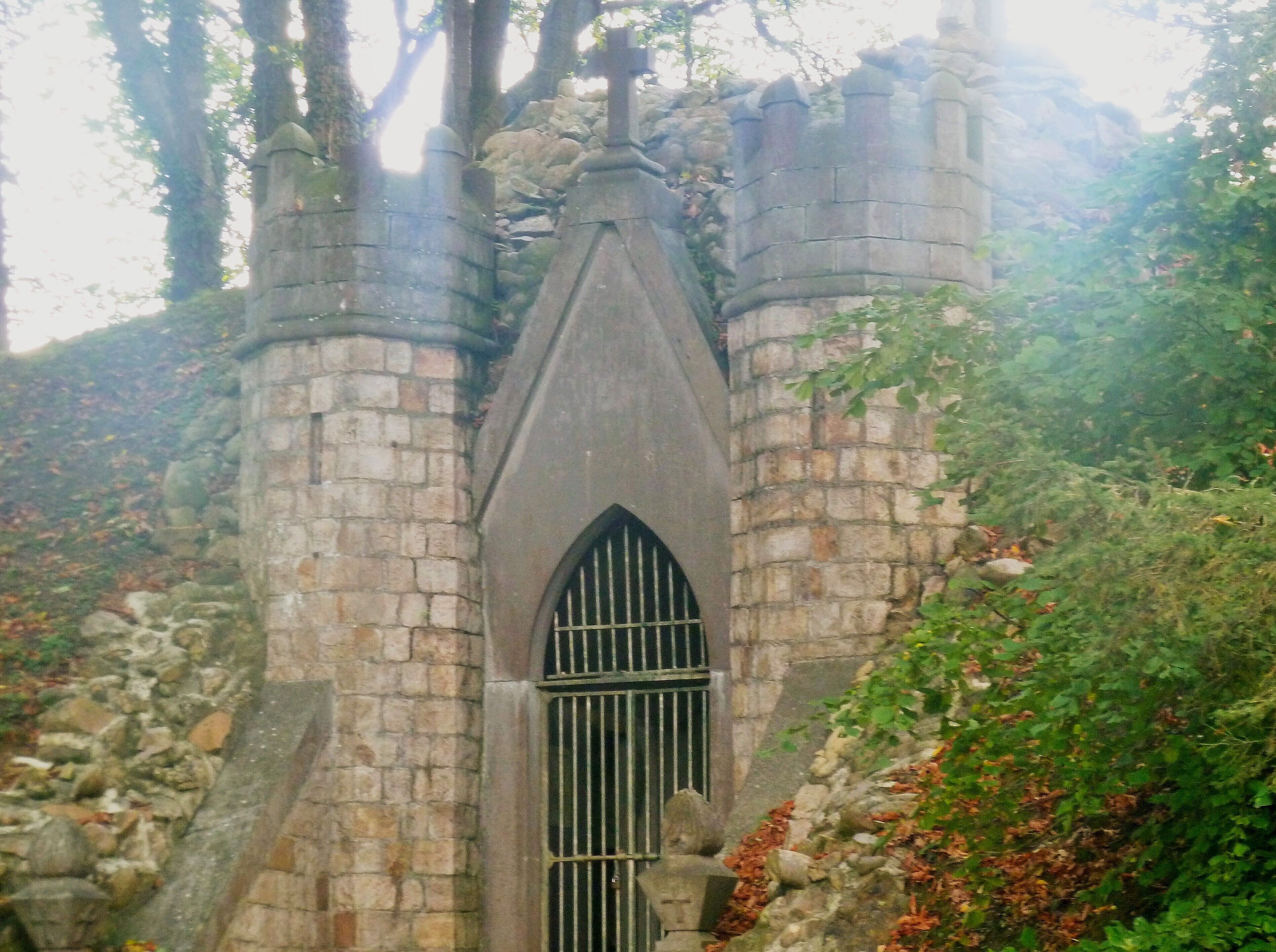 A stone tower with a pointed arch window and barred door, surrounded by trees and foliage.