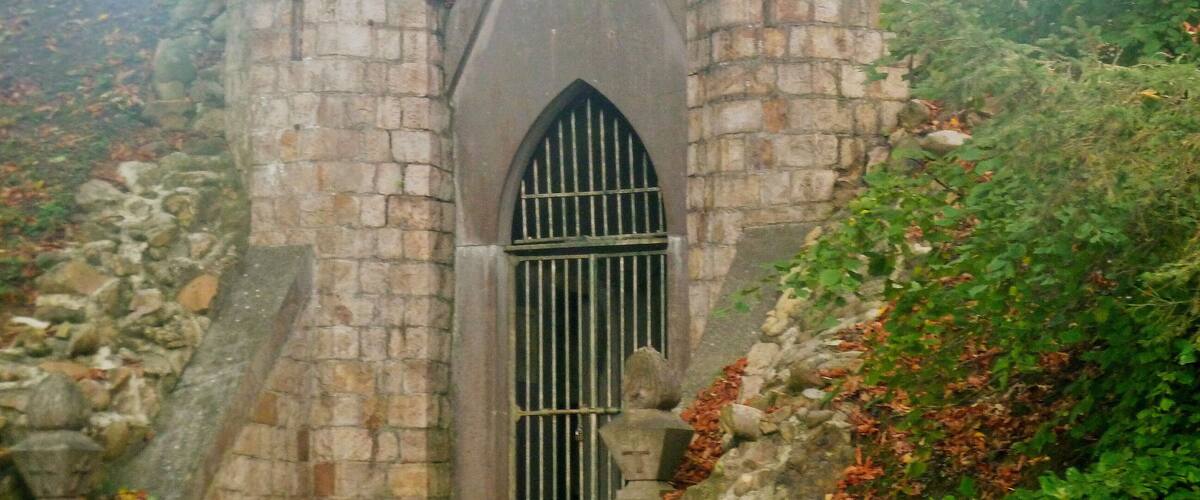 A stone tower with a pointed arch window and barred door, surrounded by trees and foliage.