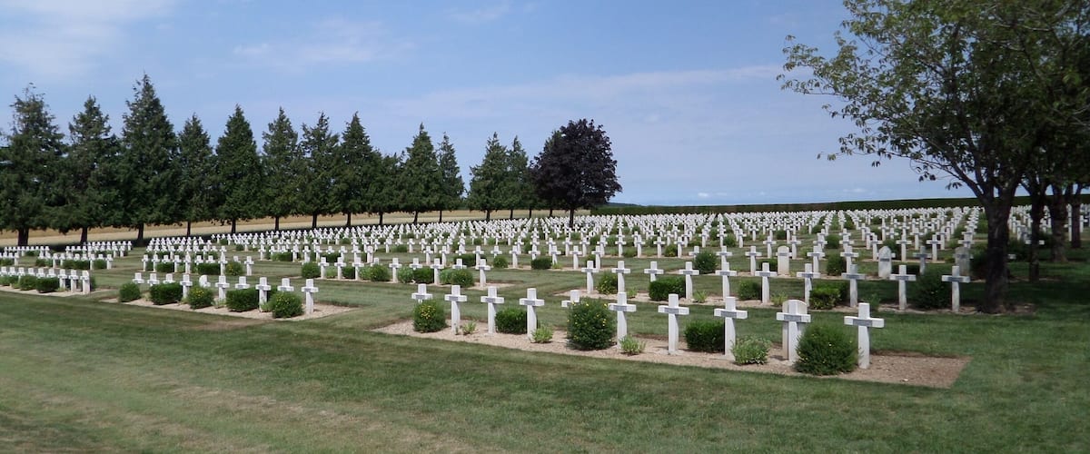 A cemetery with numerous white crosses arranged in rows, surrounded by green grass and trees.