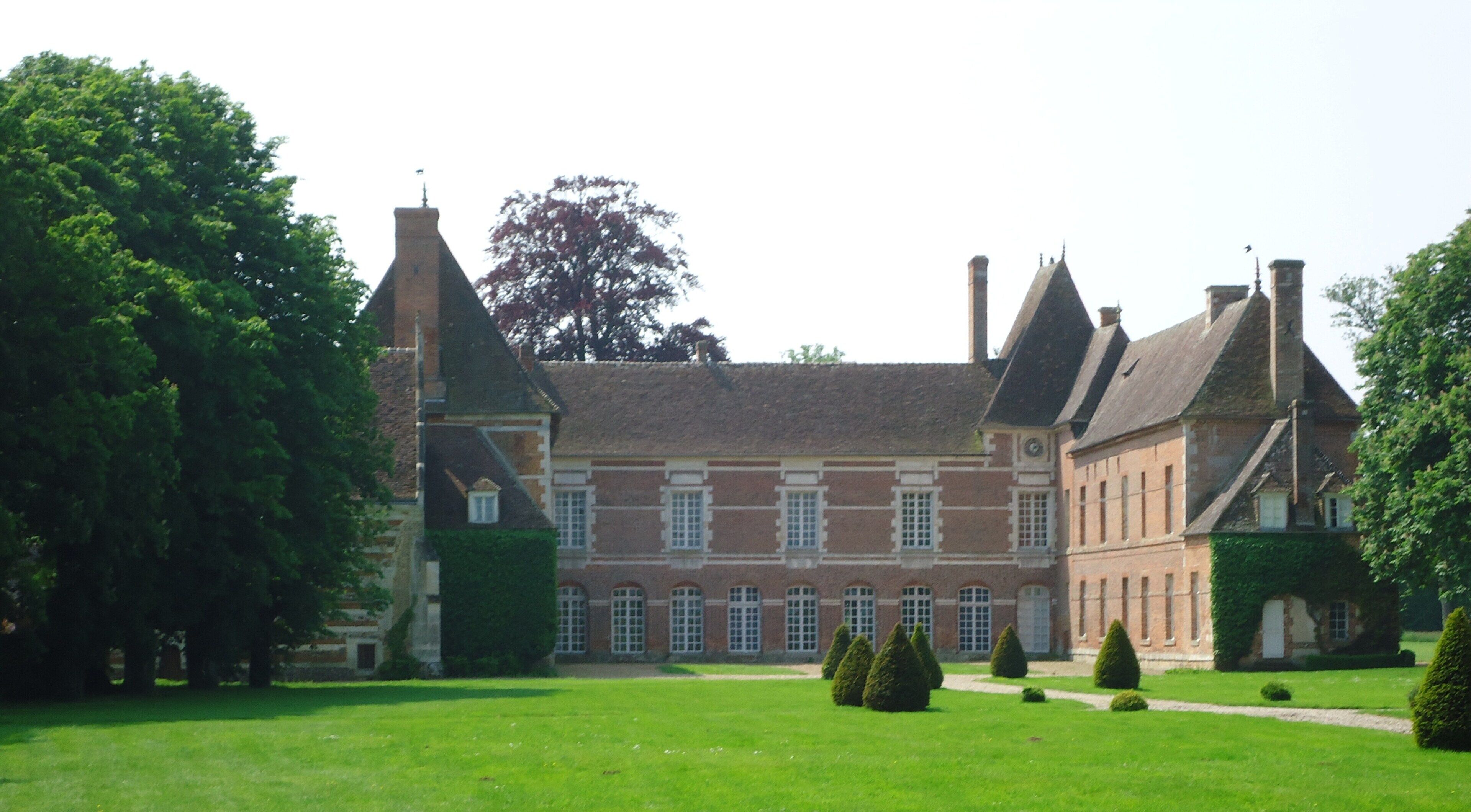 A large, historic building with a symmetrical facade, multiple chimneys, and a well-maintained garden with trimmed hedges and a lawn.