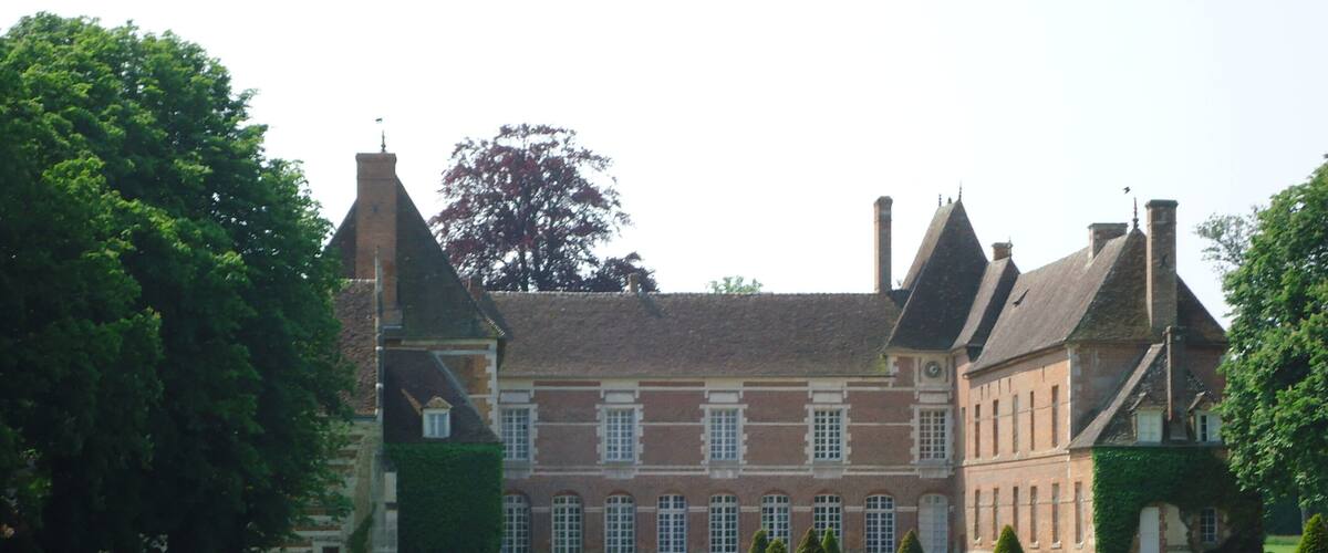 A large, historic building with a symmetrical facade, multiple chimneys, and a well-maintained garden with trimmed hedges and a lawn.