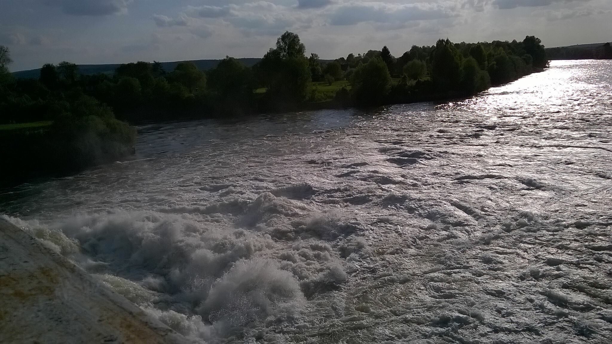 A river with turbulent water, surrounded by trees and a distant hill.