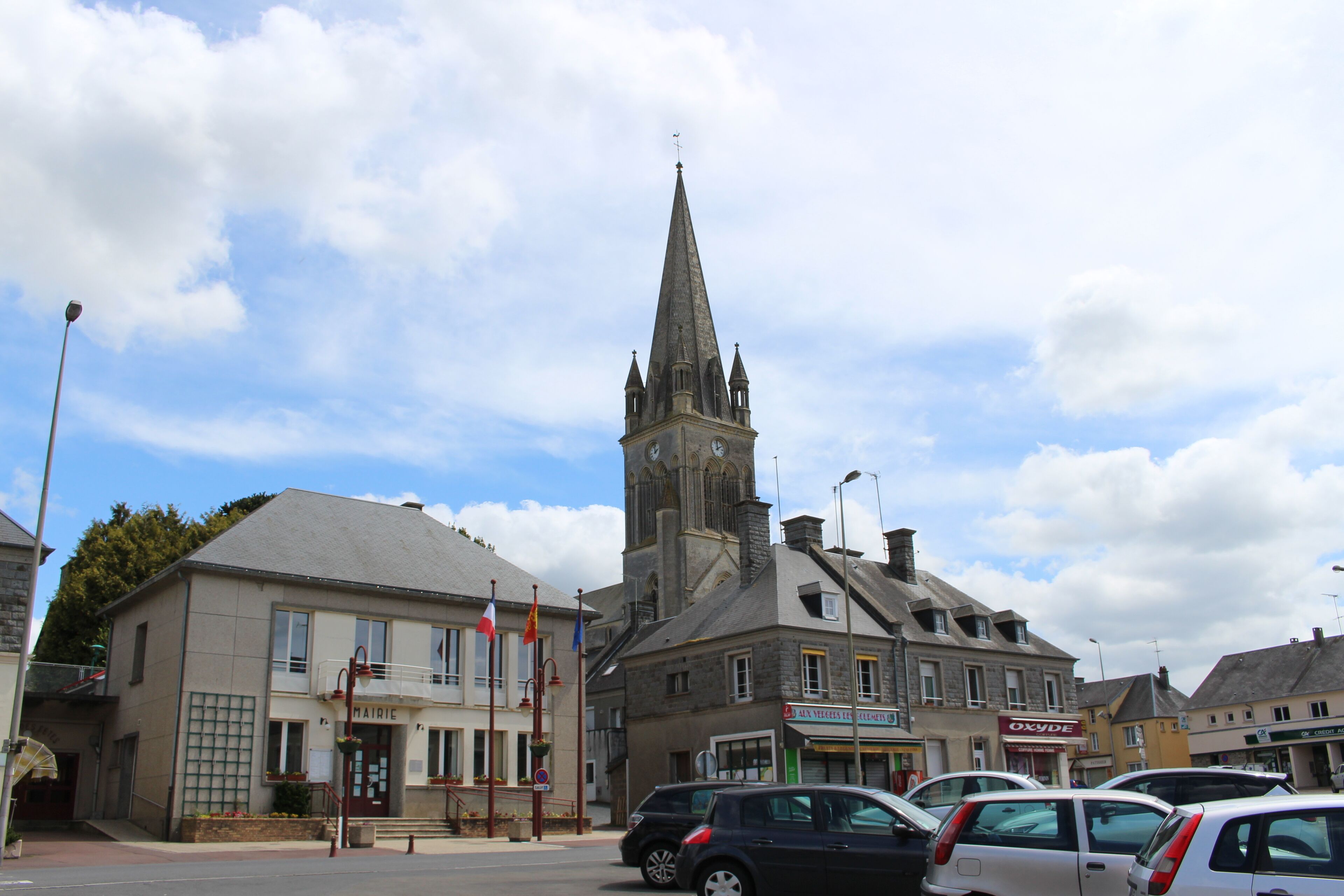 A church with a tall spire, a bakery, and parked cars in a town square.