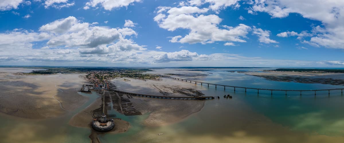 An aerial view of a coastal area with a pier extending into the water, surrounded by sandbanks and a clear sky.