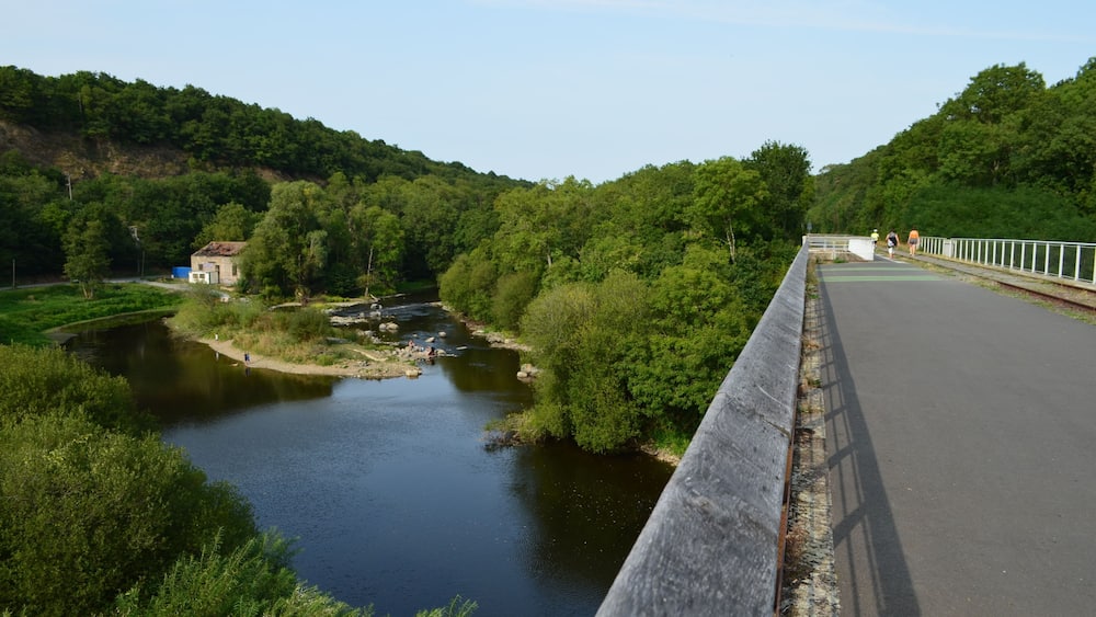 Un pont enjambant une rivière, avec un petit bâtiment sur la gauche et une végétation luxuriante de part et d’autre.