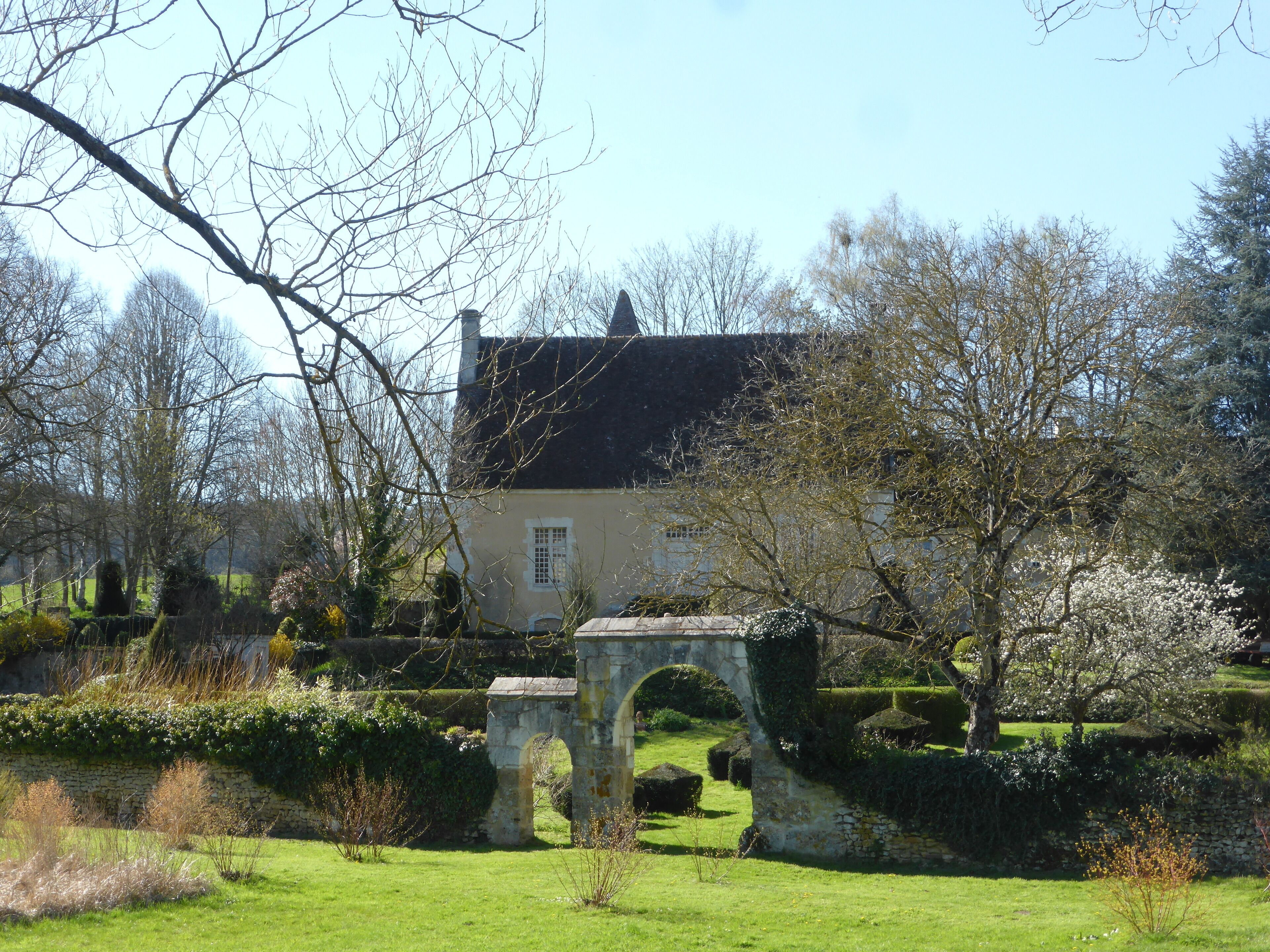 A historic stone bridge, a quaint house with a black roof, and a well-maintained garden with flowering trees.