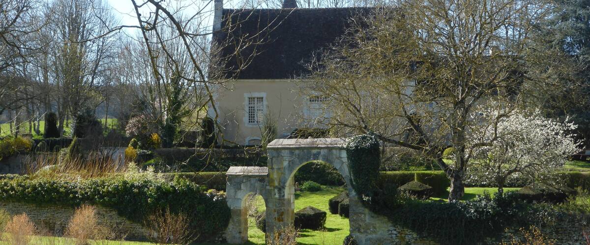 A historic stone bridge, a quaint house with a black roof, and a well-maintained garden with flowering trees.