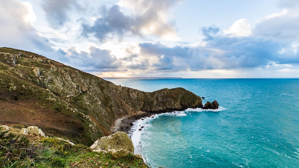 A coastal landscape with rocky cliffs, a clear blue sea, and a partly cloudy sky.