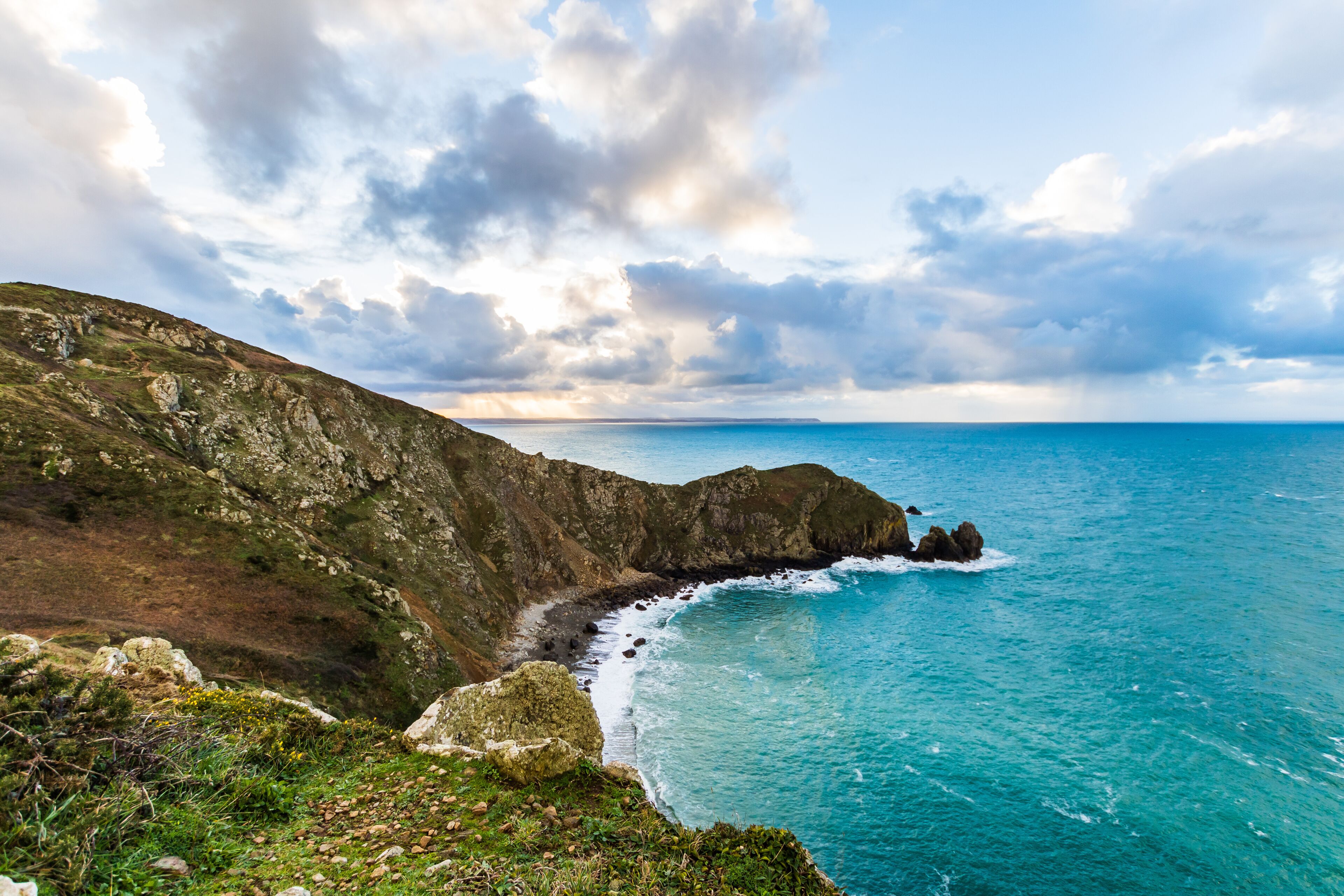 A coastal landscape with rocky cliffs, a clear blue sea, and a partly cloudy sky.