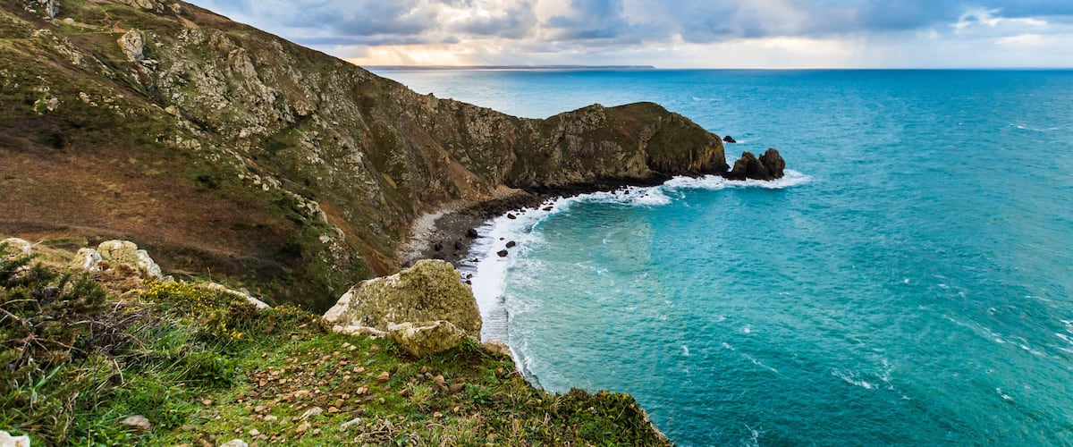 A coastal landscape with rocky cliffs, a clear blue sea, and a partly cloudy sky.