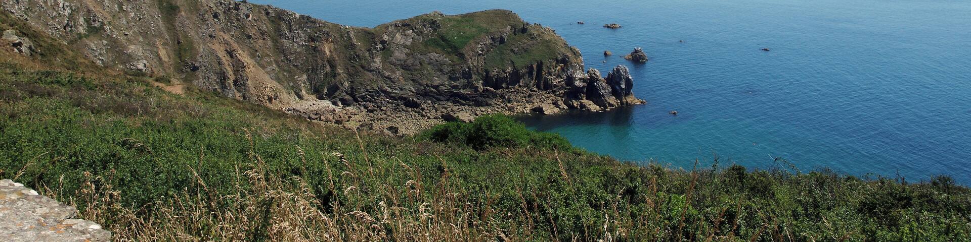 Jobourg (Manche) Le Nez de Jobourg vu depuis le Nez des Voidries. Entre les deux nez, l'Anse de Senival. Au loin, le Cap de Flamanville. "Nous dominons la mer de 128 mètres. A droite, nous avons le phare d'Auderville et le cap de la Hague ; à gauche l'anse de Vauville, que ferme dans le lointain, à une quinzaine de kilomètres, le cap de Flamanville. Une légère brume en haute mer nous cache les îles anglo-normandes. Tout près, au sud, s'allonge le Nez de Jobourg, moins haut mais plus déchiqueté que la pointe sur laquelle nous sommes. Là, finit le passage de la déroute et commence le Raz-Blanchard. Dans les hautes falaises de Jobourg existent des grottes d'un accès difficile : on y descend en suivant un sentier périlleux établi par le T. C. F. Si dangereux que soient ces parages pour la navigation il n'en ont pas moins été fréquentés par les fraudeurs. Il y avait autrefois dans la région des sociétés organisées pour la fraude du tabac. La configuration de la côte, riche en cachettes, facilitait ce trafic. Les fraudeurs, les «hagards», selon l'expression locale, sont devenus plus rares. Les lourdes amendes et la prison qui les menacent aujourd'hui ont presque entièrement arrêté leur fructueux commerce." Compte rendu de l'excursion faite par la Société Historique et Archéologique de l'Orne dans le Cotentin 29 Août — 2 Septembre 1927 - Caen, 26 janvier 1928. Camille de Mons..