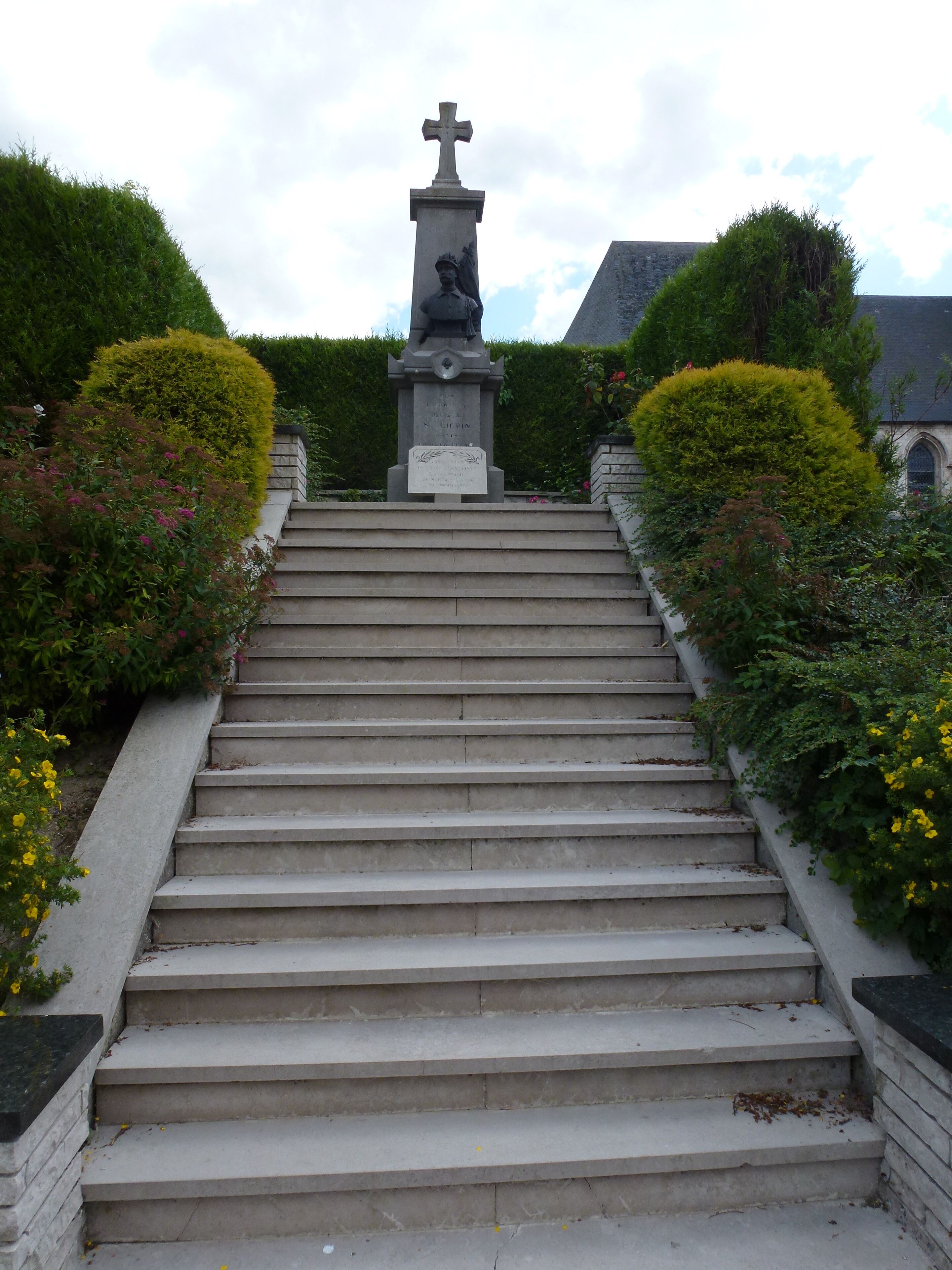 A set of stone steps leading up to a statue with a cross on top, surrounded by well-maintained hedges and flowering plants.