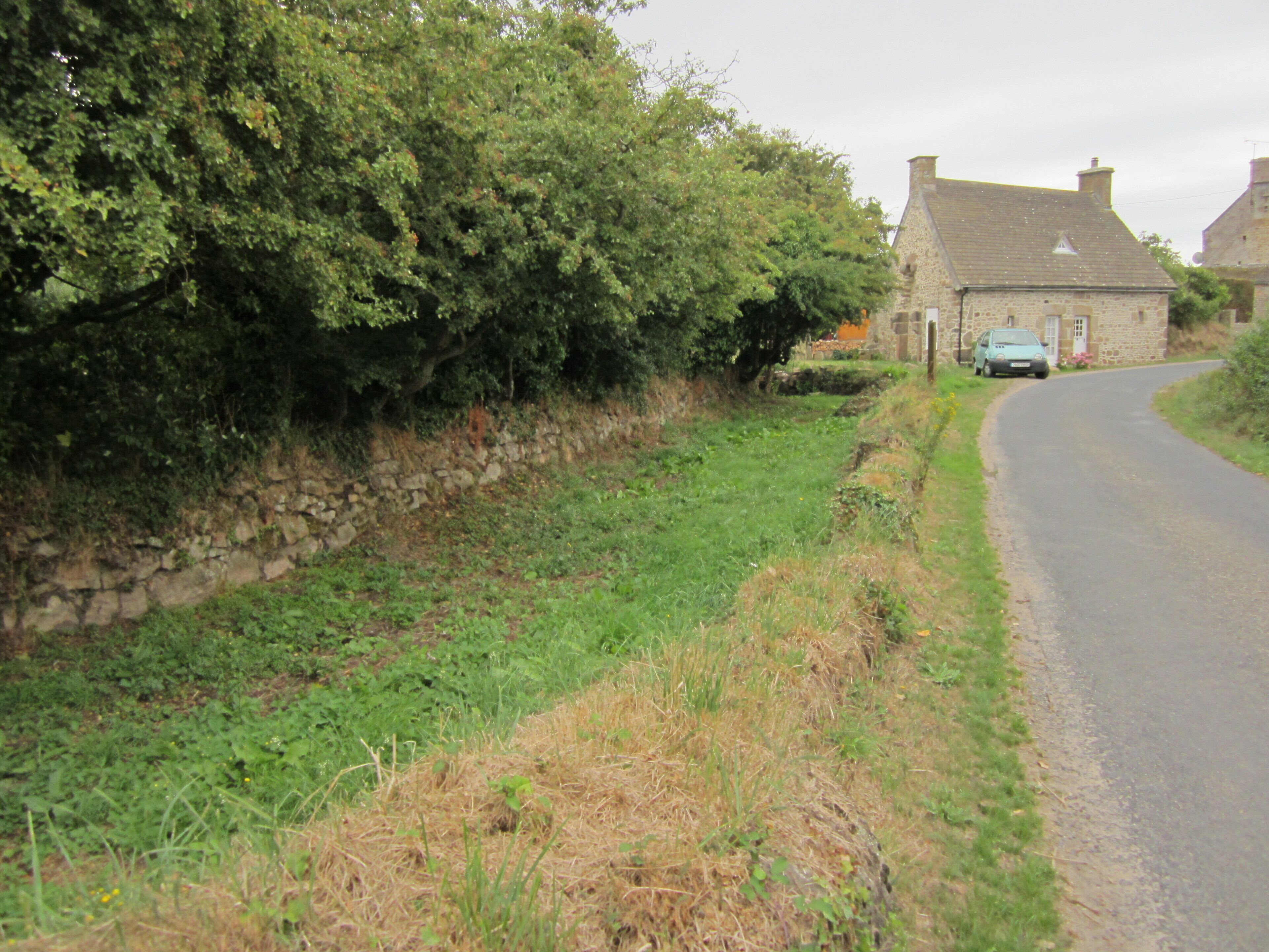 A rural road with a stone wall, greenery, and a stone house.