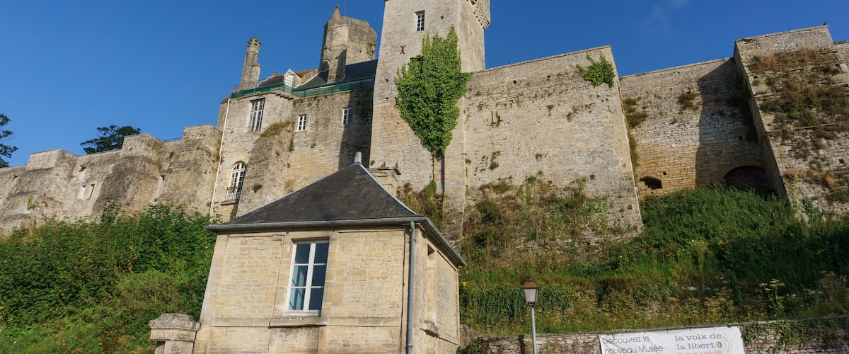 A historic stone building with a tower, surrounded by greenery and a clear blue sky.