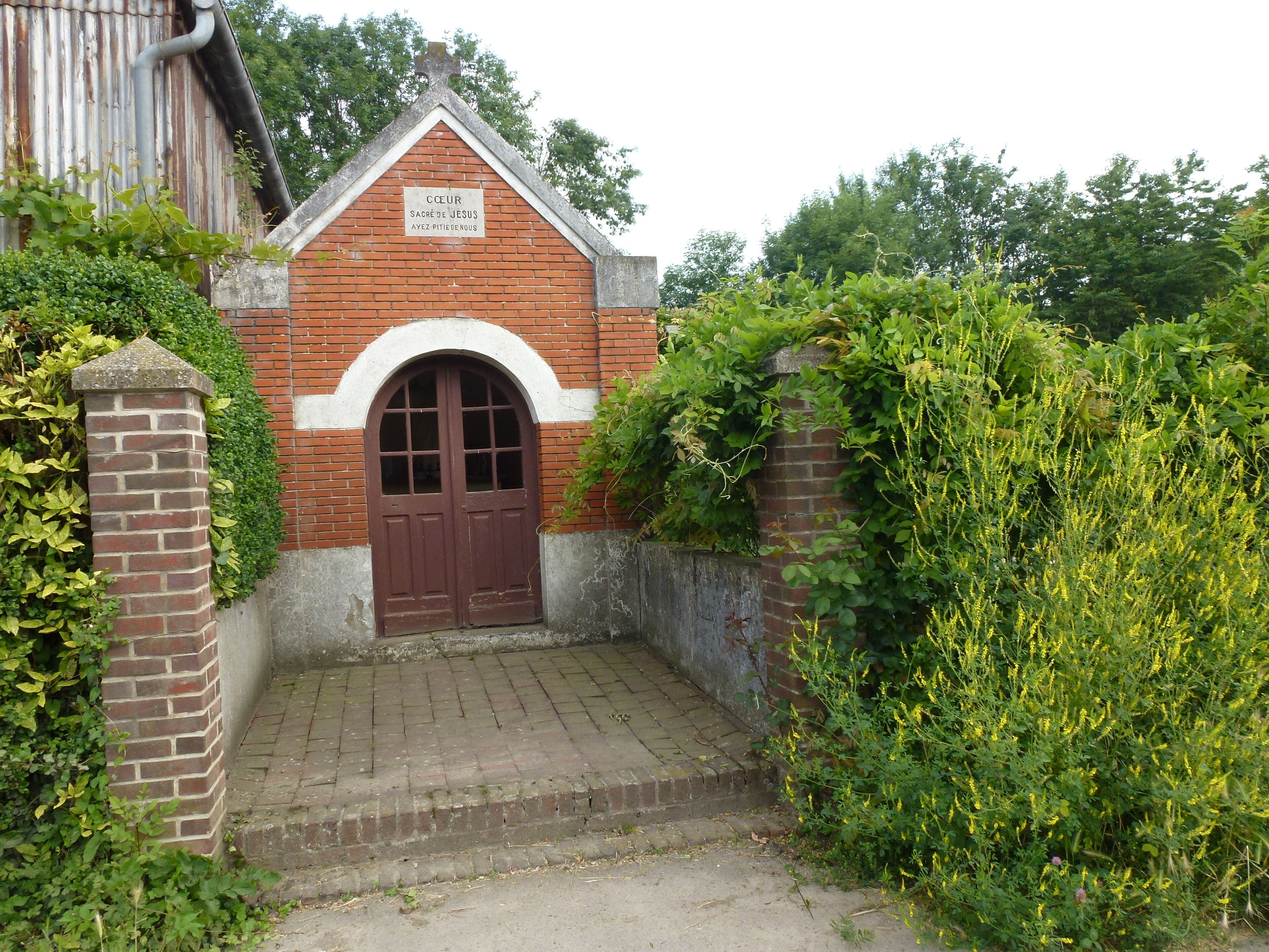 A small brick building with a red door and a sign reading "Cœur Sacré de Jésus Ayez Pitié envers nous".