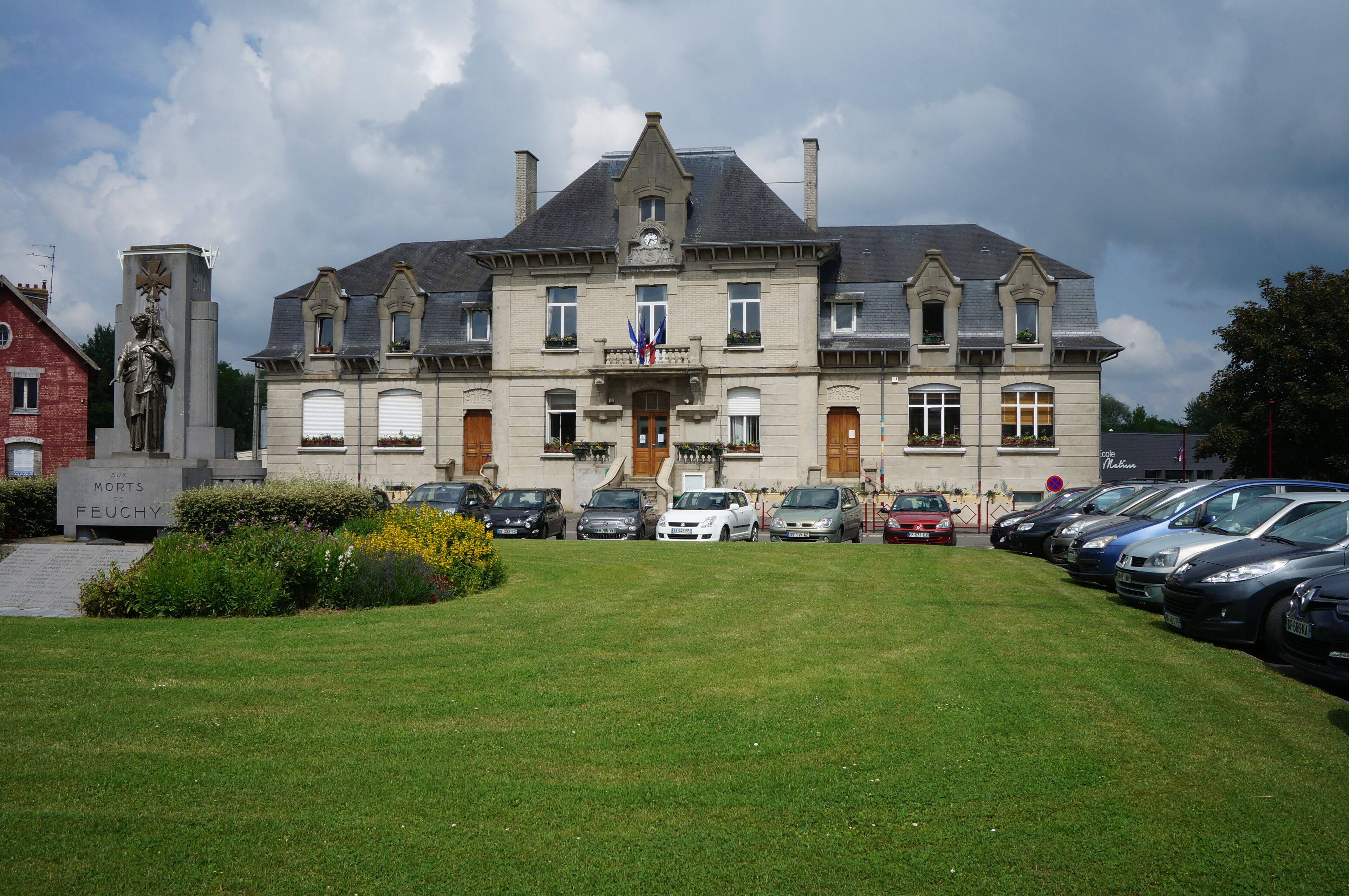 A large, historic building with a statue in front, surrounded by a well-maintained lawn and parked cars.