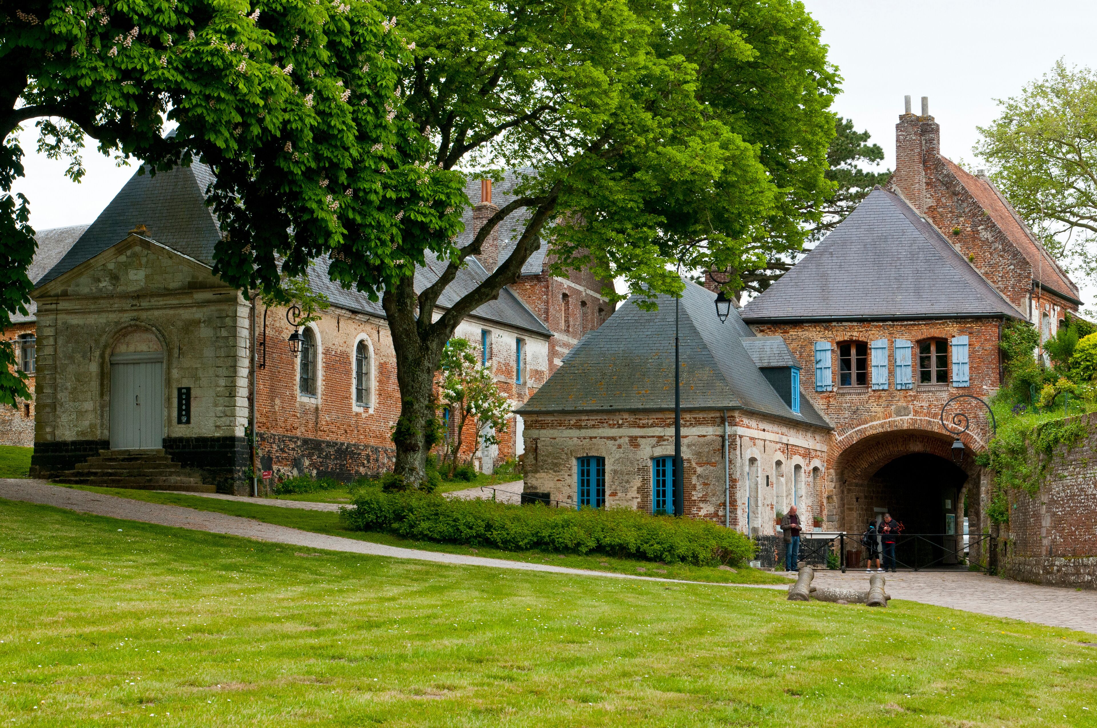 A historic building with a stone facade, a large tree in front, and a brick archway.