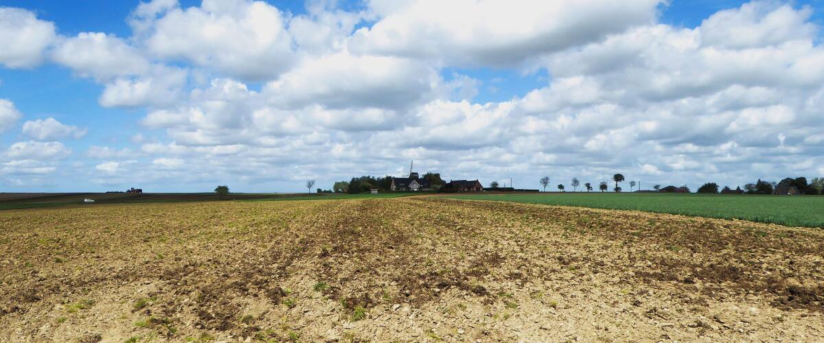 A plowed field with a farmhouse in the distance under a partly cloudy sky.