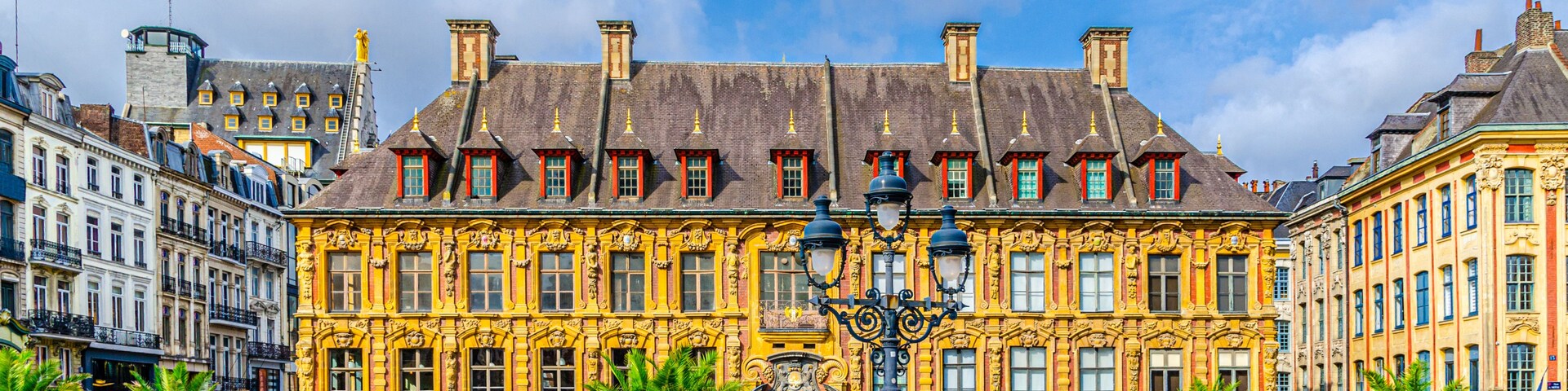 Un bâtiment historique aux façades jaunes richement décorées et aux encadrements de fenêtres rouges, entouré de rues pavées et de palmiers.