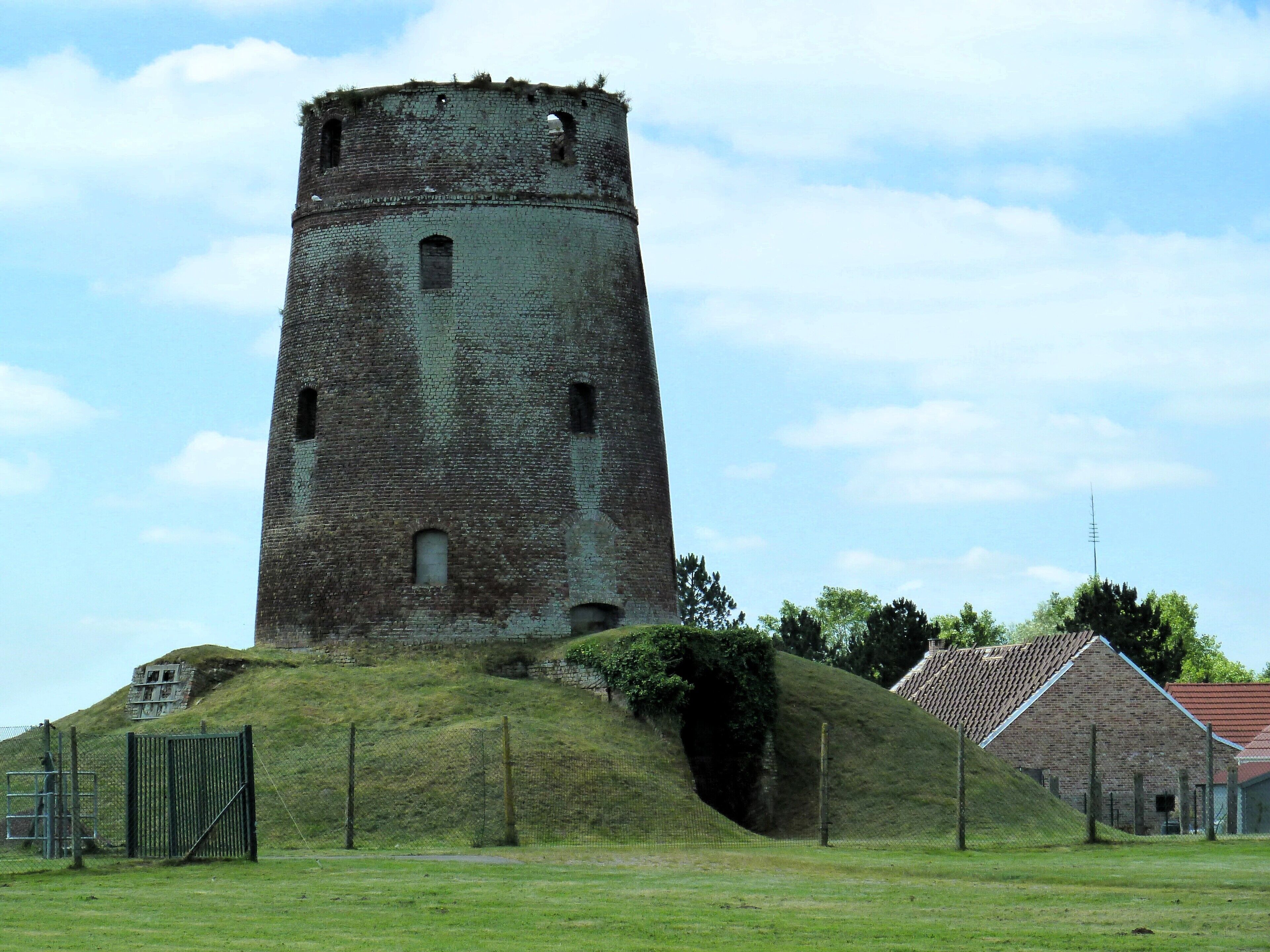 A historic stone tower with a conical roof, surrounded by a grassy area and a small building with a red roof.