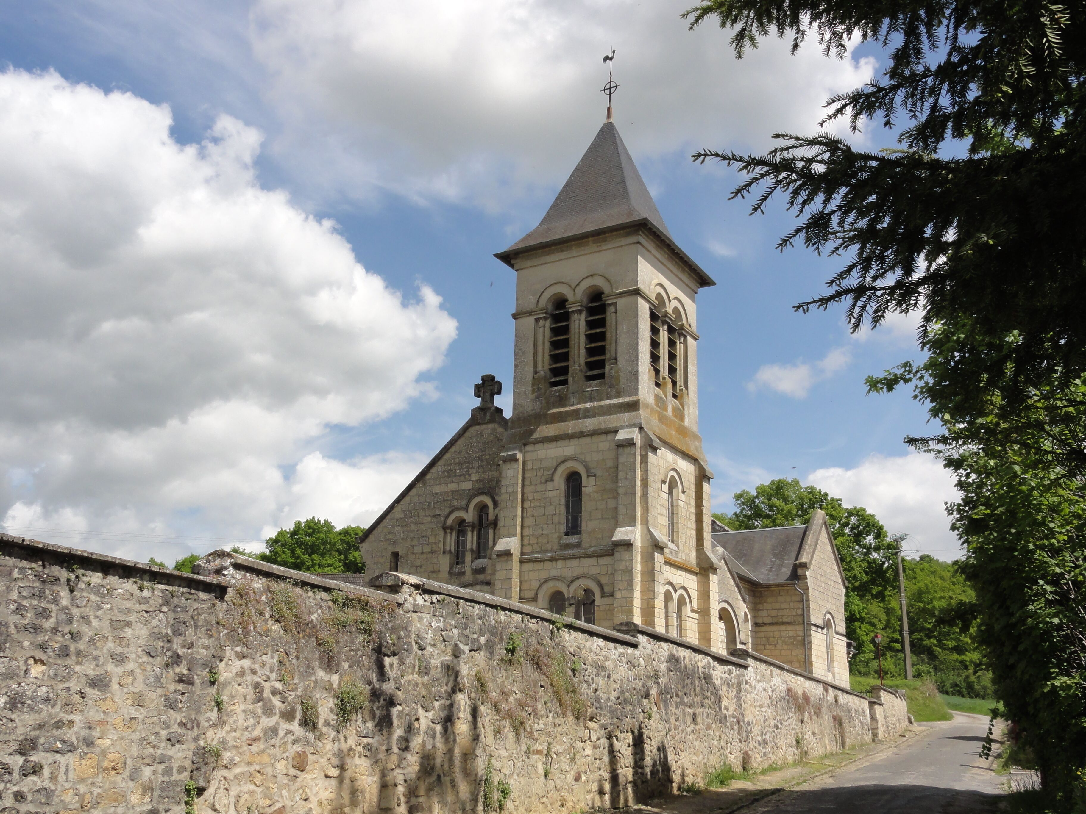A historic stone church with a tall bell tower and a weather vane, surrounded by a stone wall and trees.